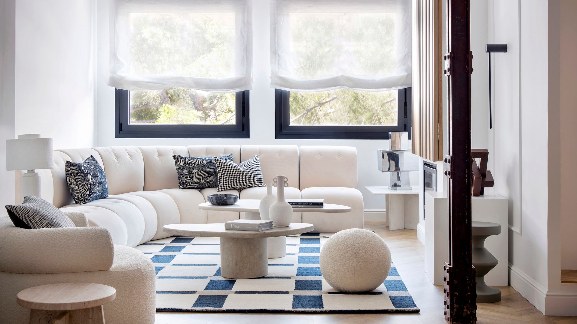 Salón con alfombra bicolor con motivos geométricos, sofá blanco curvo, ventanas con el marco negro, estores de color blanco, mesas de centro, cojines sobre el sofá, puff redondo 
