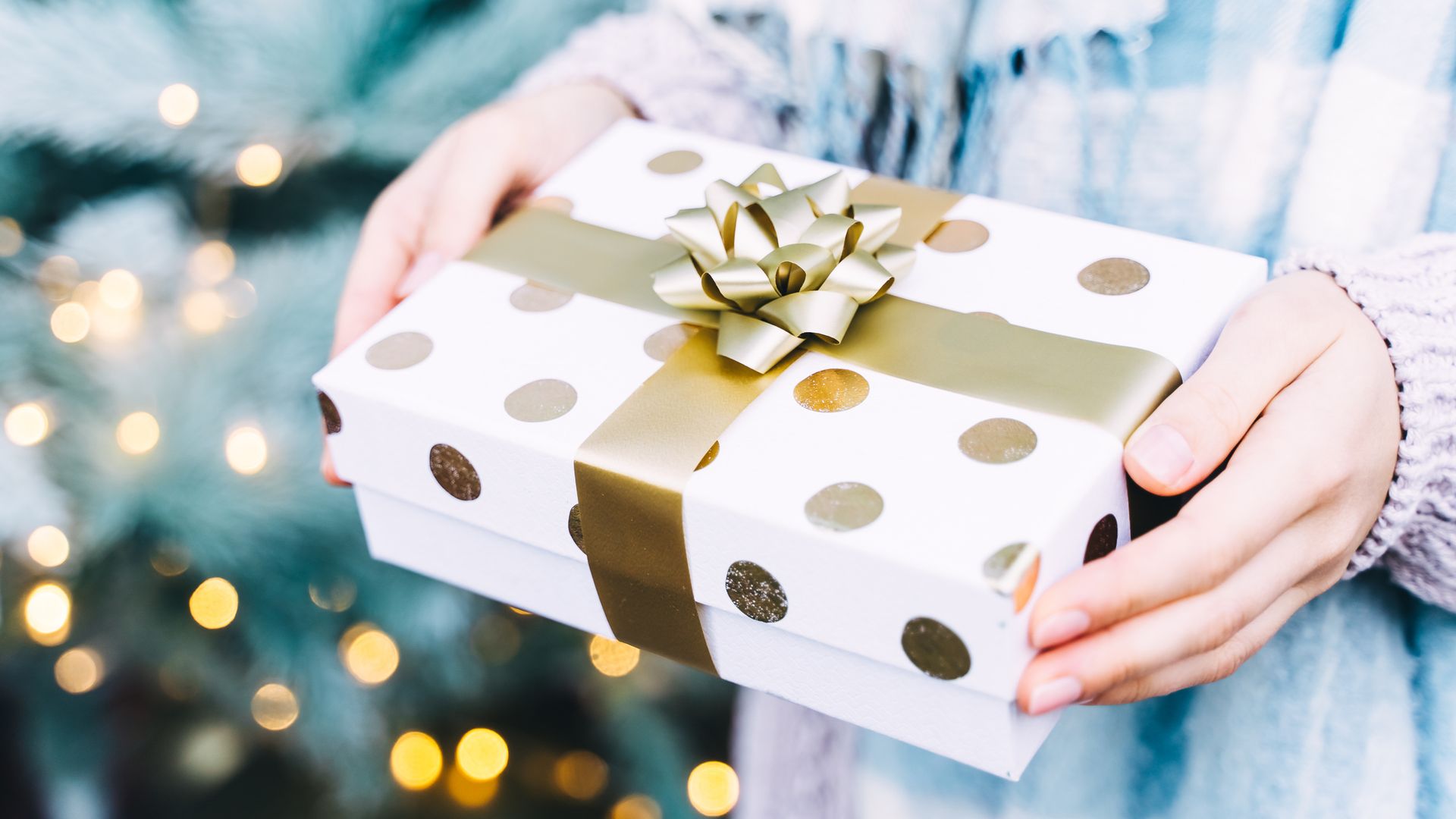 Mujer con un regalo de Navidad entre las manos envuelto en blanco y dorado. Lleva una bufanda azul y detrás hay un abeto iluminado.