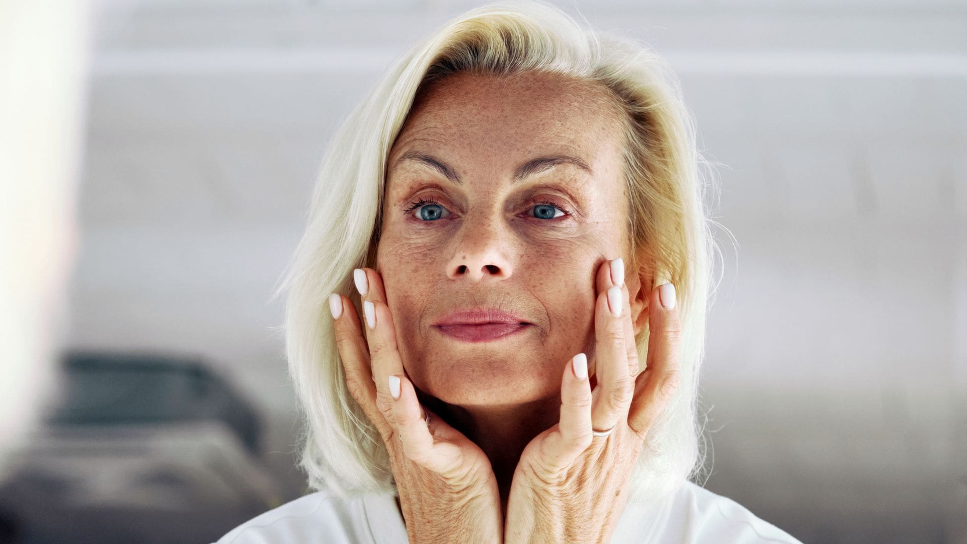 Rostro de mujer madura con pelo rubio claro y ojos azules tensándose la piel con las manos