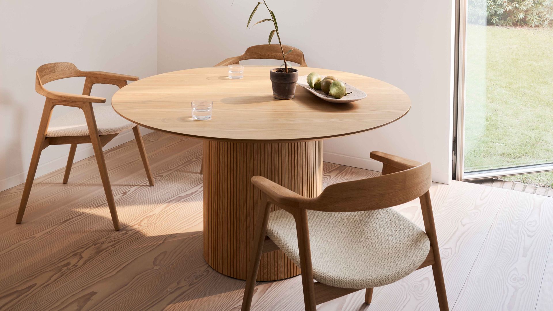 Comedor con mesa redonda de madera, sillas de madera con brazos y asiento tapizado, salida al jardín y maceta con planta