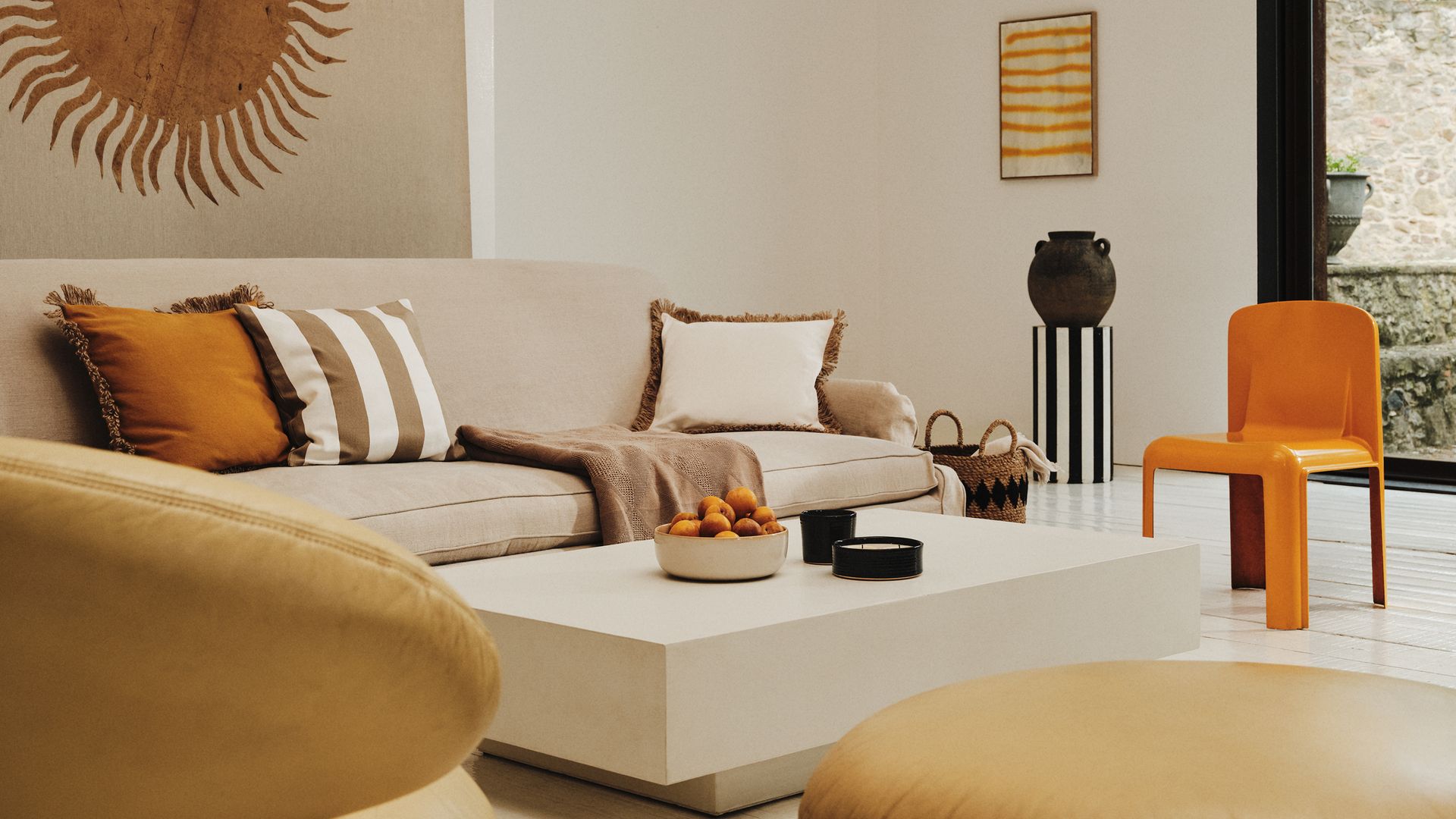 Salón con tapiz en la pared del sofá, sofá beige, mesa de centro blanca, adornos sobre la mesa de centro, cojines de rayas y lusos, silla naranja, cuadro en la pared, pedestal de rayas con vasija