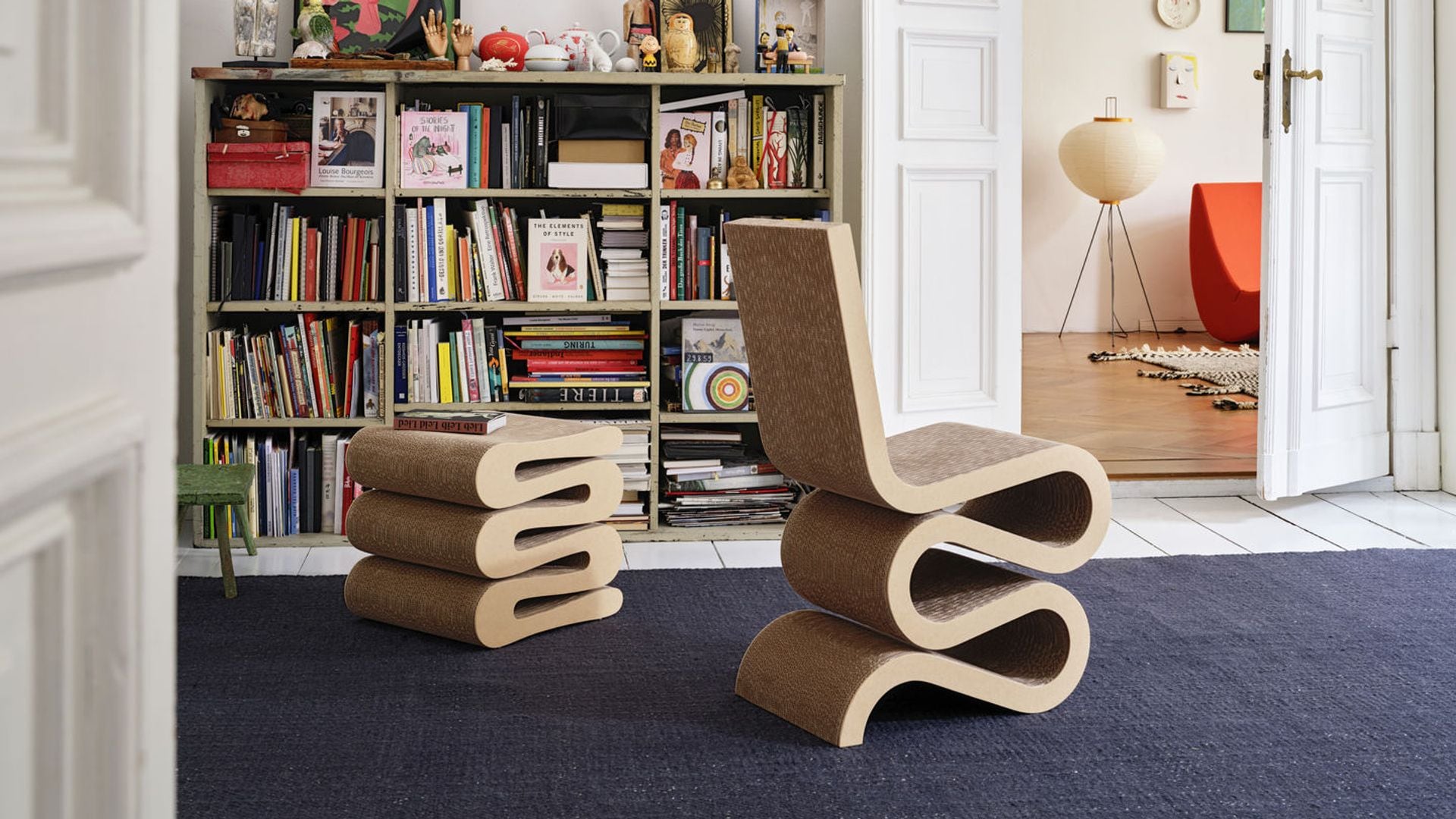 Silla y mesa auxiliar de Frank Gehry en cartón, alfombra azul, librería baja con libros