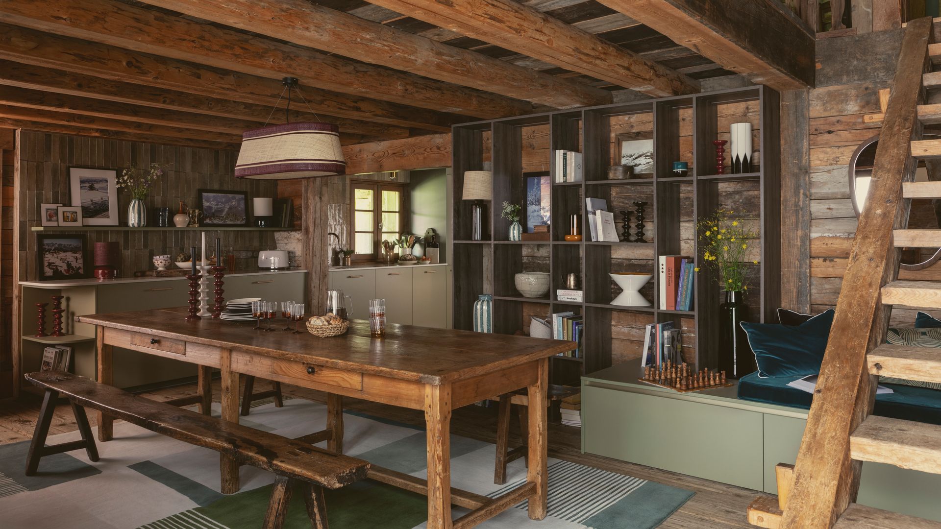 Cabaña con comedor con mesa y banco de madera, muebles verdes, estantería, lámpara de techo, escaleras