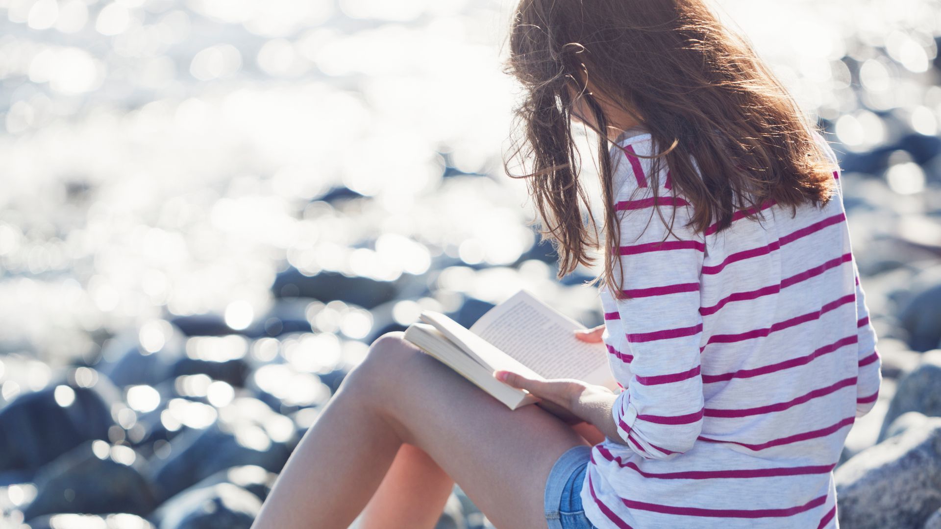 Chica adolescente leyendo un libro en una playa con ropa de verano, sobre una playa de rocas.