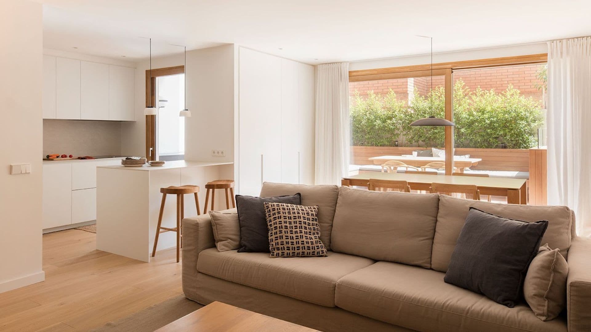 Cocina abierta al salón y al comedor, con salida al jardín, cocina blanca, con península con taburetes, muebles de suelo a techo sin tiradores, sofá marrón con cojines, mesa de comedor de madera, lámpara de techo negra