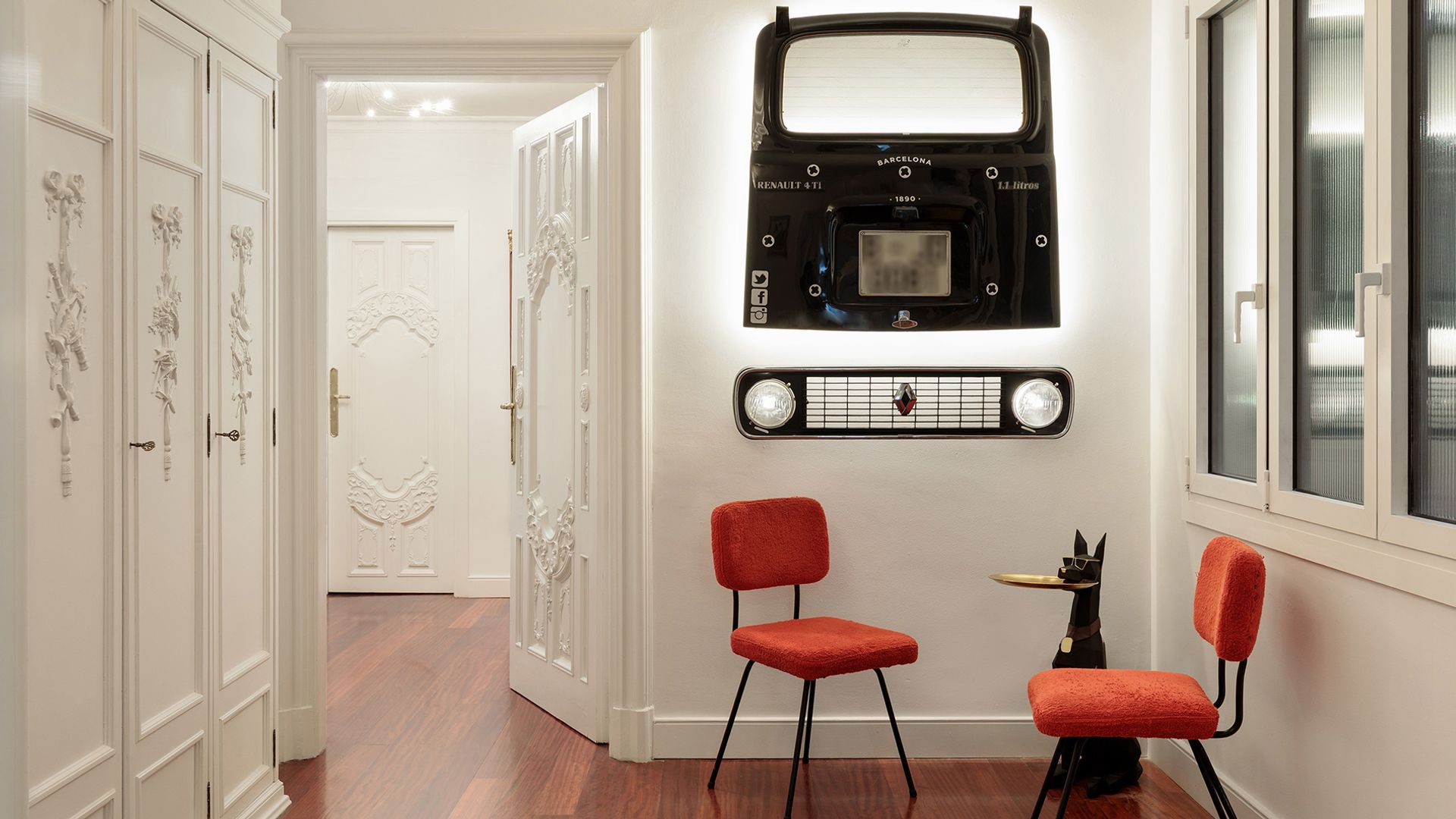 Recibidor con dos sillas rojas, una mesa auxiliar en forma de gato, una lámpara de pared en forma de coche antiguo, suelo de madera oscuro