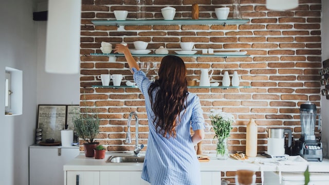 Mujer en el fregadero de la cocina, de espaldas, colocando unas tazas en las estanterías que tiene en la pared sobre la encimera.