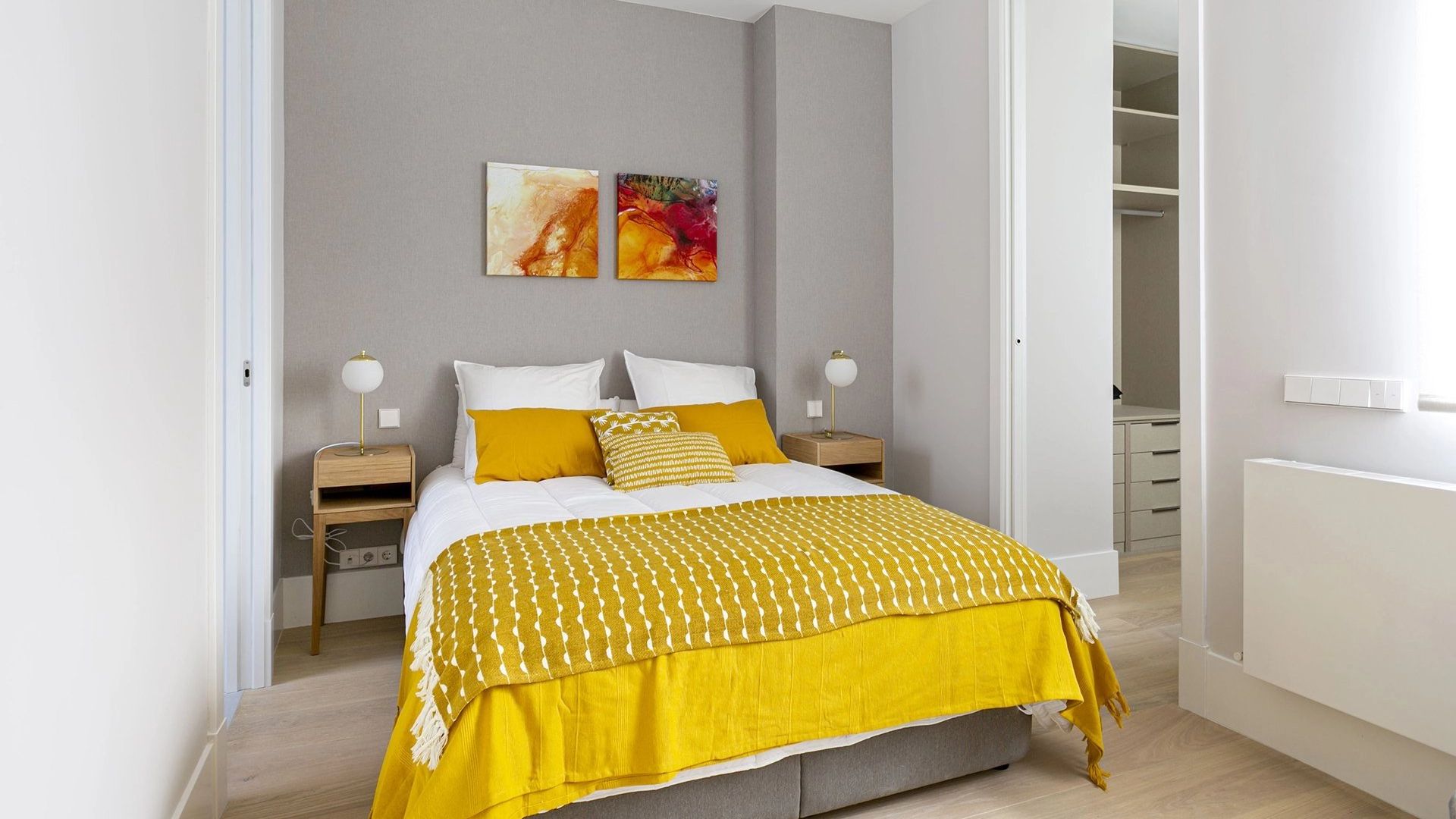 Dormitorio con pared del cabecero gris, ropa de cama amarilla, cuadros en la pared del cabecero, radiador 