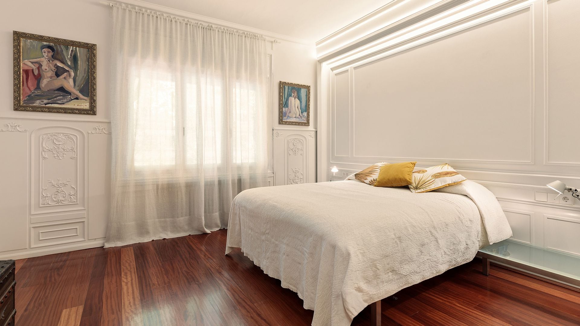 Dormitorio con paredes con molduras blancas, suelo de madera oscura, ropa de cama blanca, cortinas blancas, cuadros en la pared