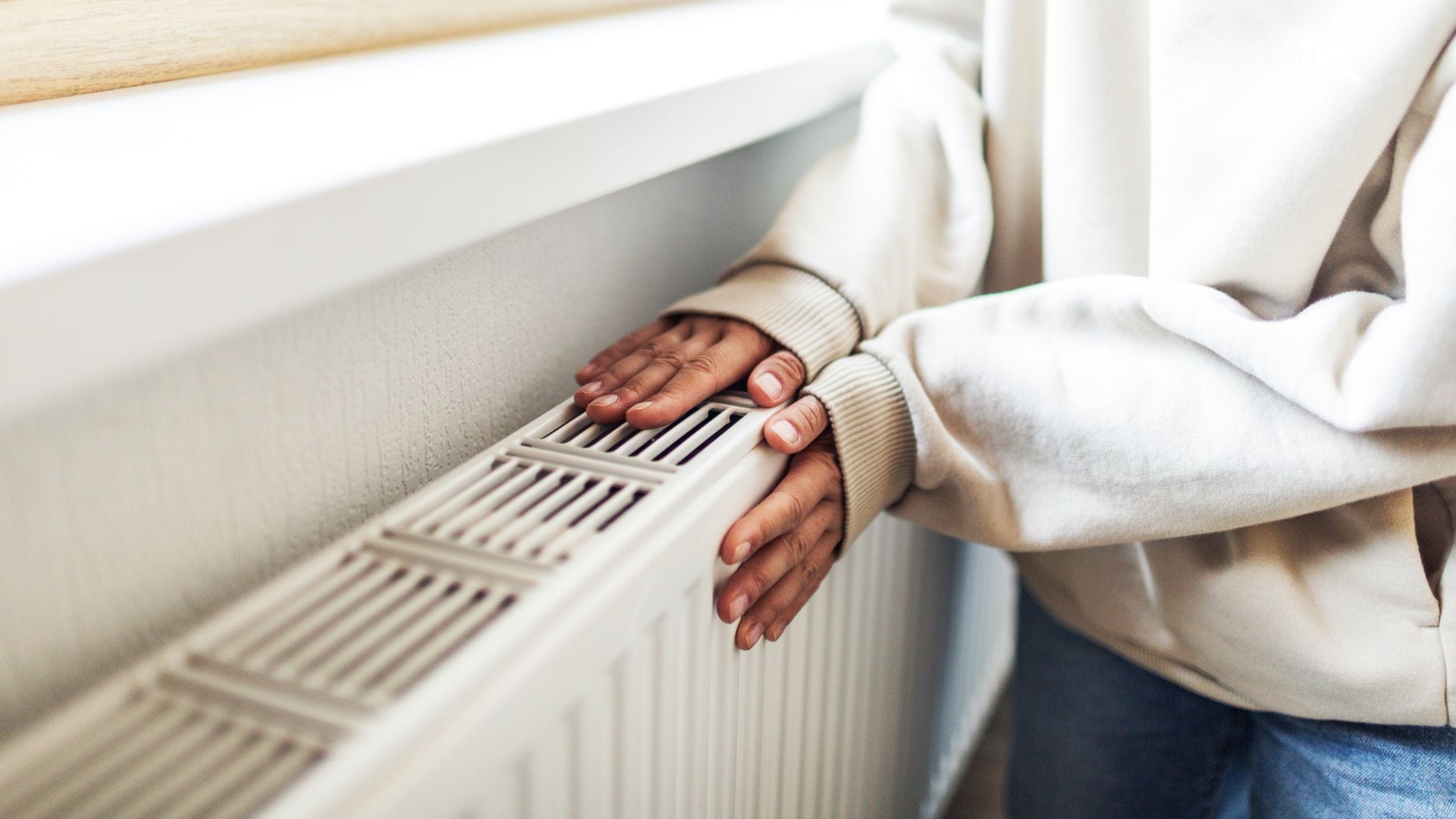 Manos de mujer tocando un radiador blanco, con jersey 'beige' y pantalón vaquero. Es un plano detalle.