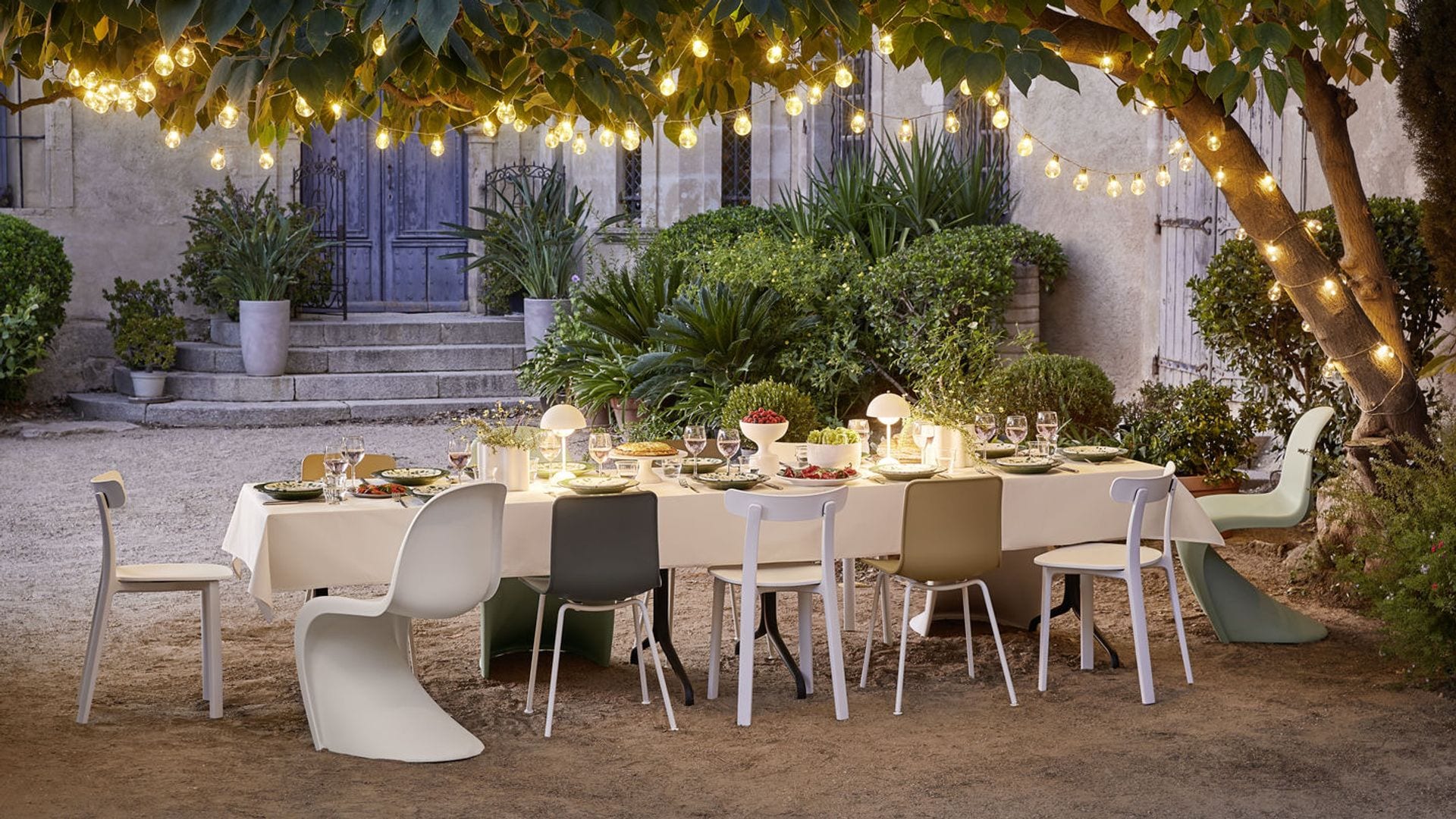 Mesa de verano en jardín bajo árbol con guirnalda de luces, sillas Panton, mantel blanco, lámparas de sobremesa blancas y rojas