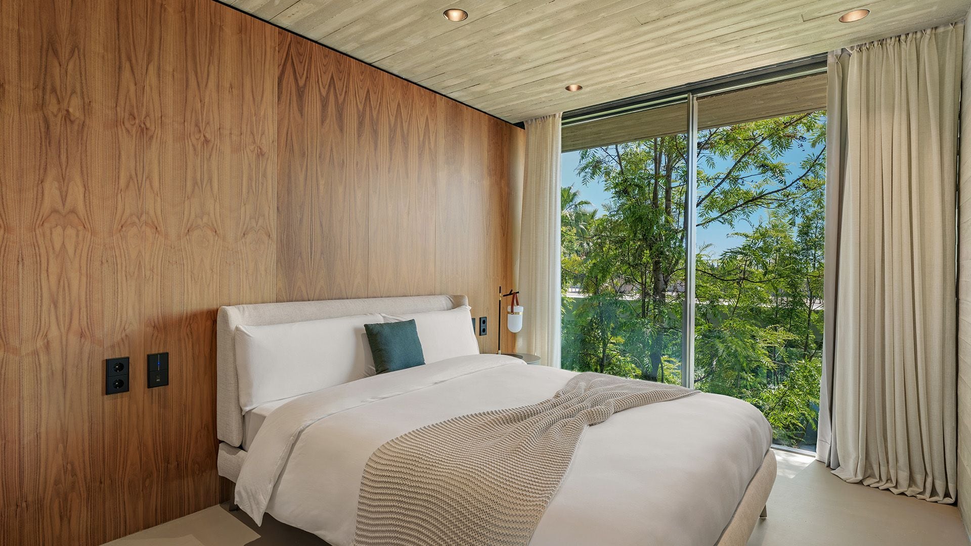 Dormitorio con pared de madera, cama con cabecero tapizado, ropa de cama blanca, cortinas beiges y gran ventanal con vistas al jardín