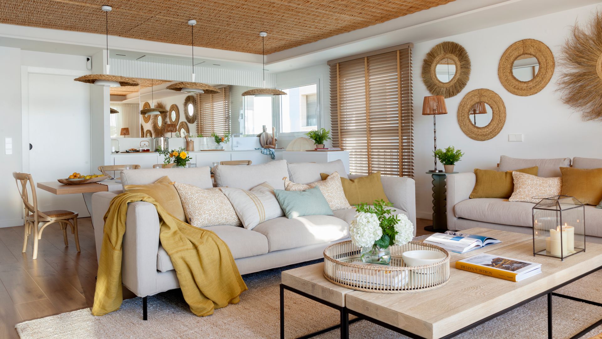 Salón comedor con aire mediterráneo, con techo de fibras vegetales, espejos redondos en la pared del sofá con el marco de fibras vegetales, lámparas de techo de fibras vegetales sobre la mesa de comedor, mesa de centro de madera con estructura metálica negra, decorada con velas y jarrones con flores, sofás beiges