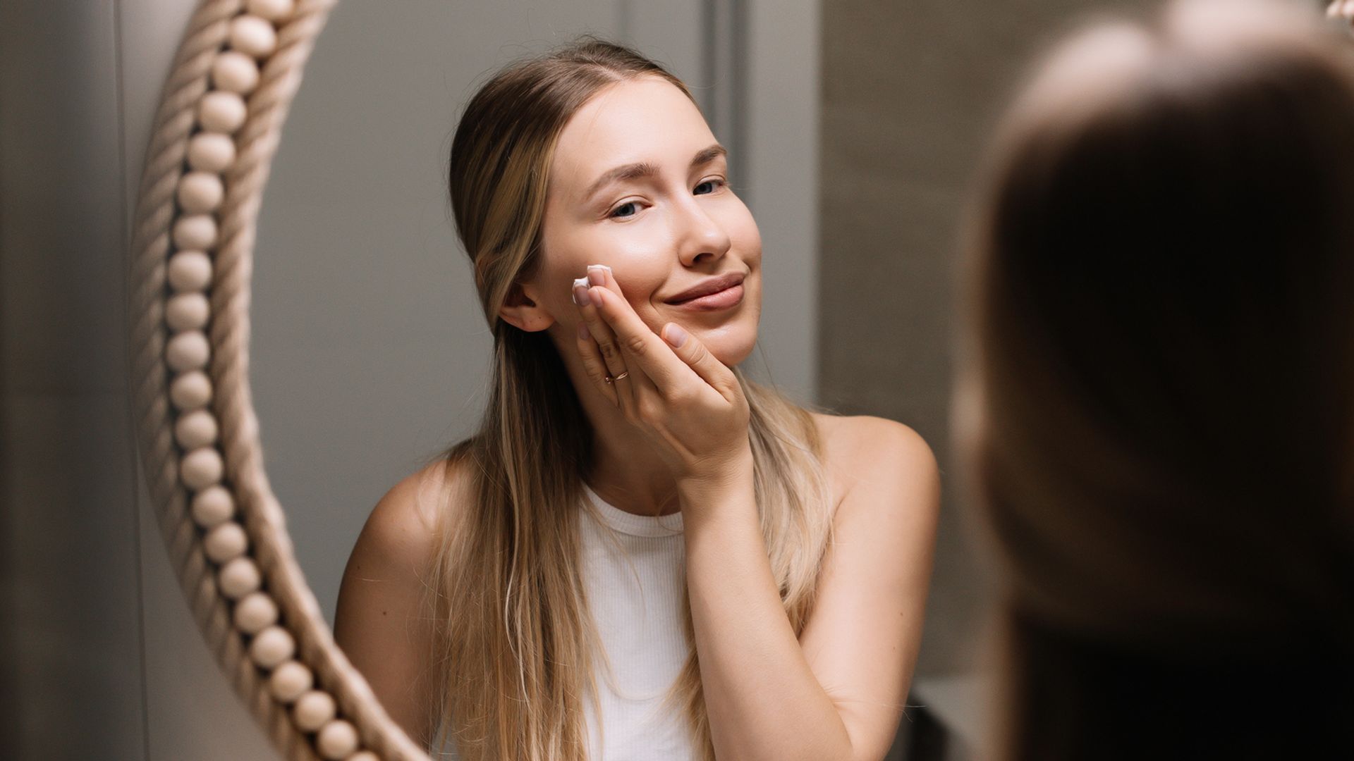 Mujer joven rubia frente al espejo de su cuarto de baño aplicándose crema en el rostro