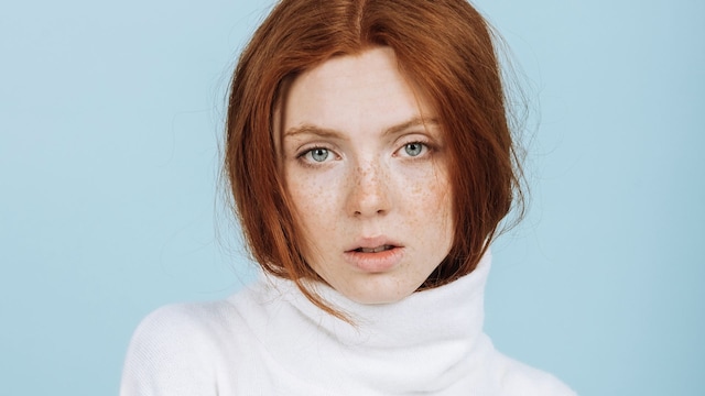 Mujer pelirroja de ojos claros y cabello bonito con jersey de cuello alto mirando a la cámara en primer plano con fondo azul