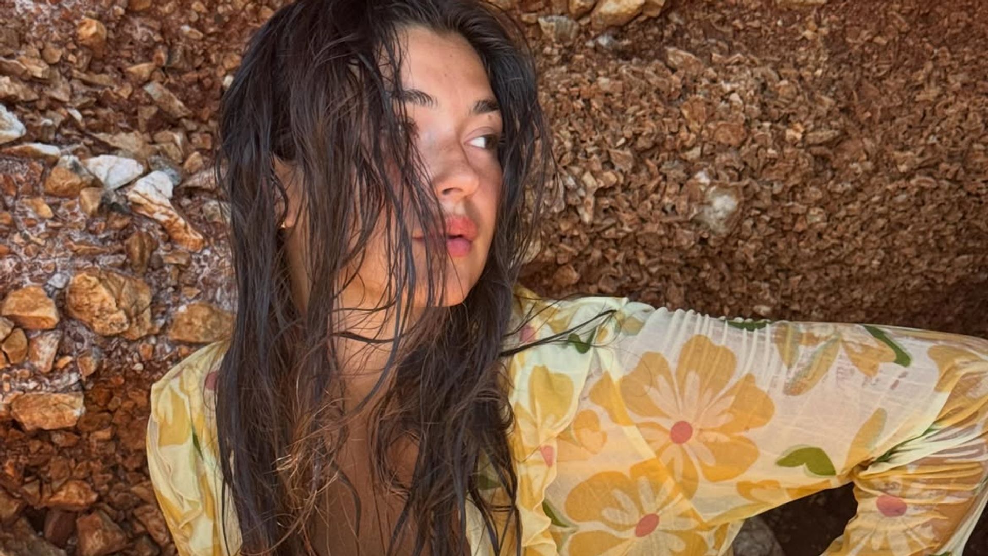 Mujer con el pelo largo y mojado descansa al sol junto a unas rocas.
