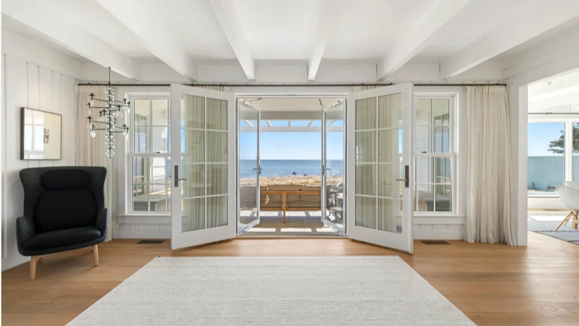 Salón con suelo de madera y salida al porche con vistas al mar, cortinas blancas, vigas techo