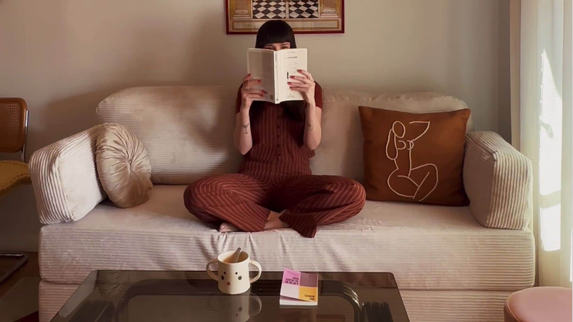 Mujer sentada leyendo un libro en sofá beige con cojines mostaza y de raso crema, mesa de centro de cristal ahumado, con taza y libro