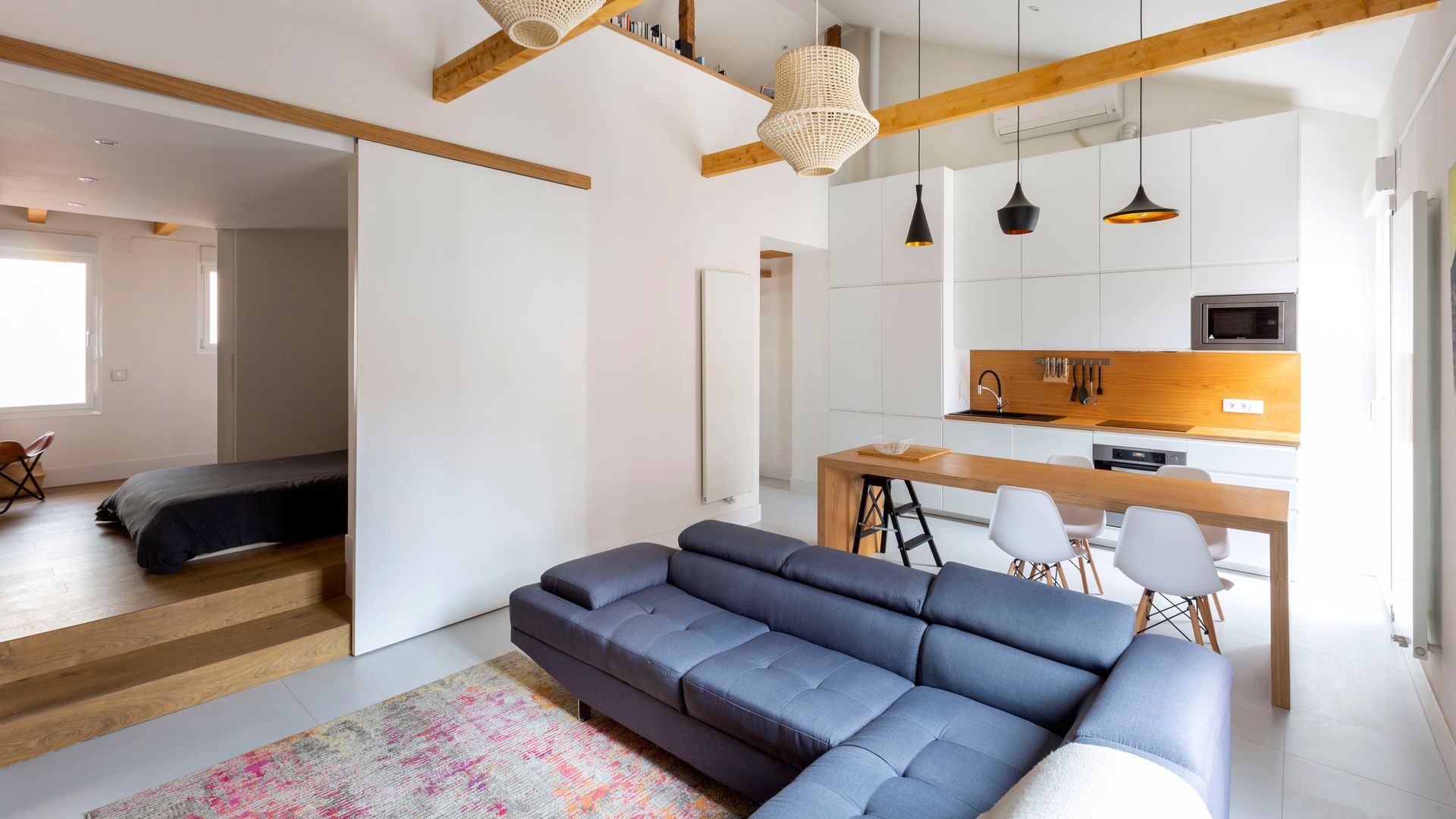 Salón comedor de estilo nórdico con dormitorio conectado con puerta corredera blanca, lámparas de techo, cocina blanca con el antepecho de madera y sofá gris