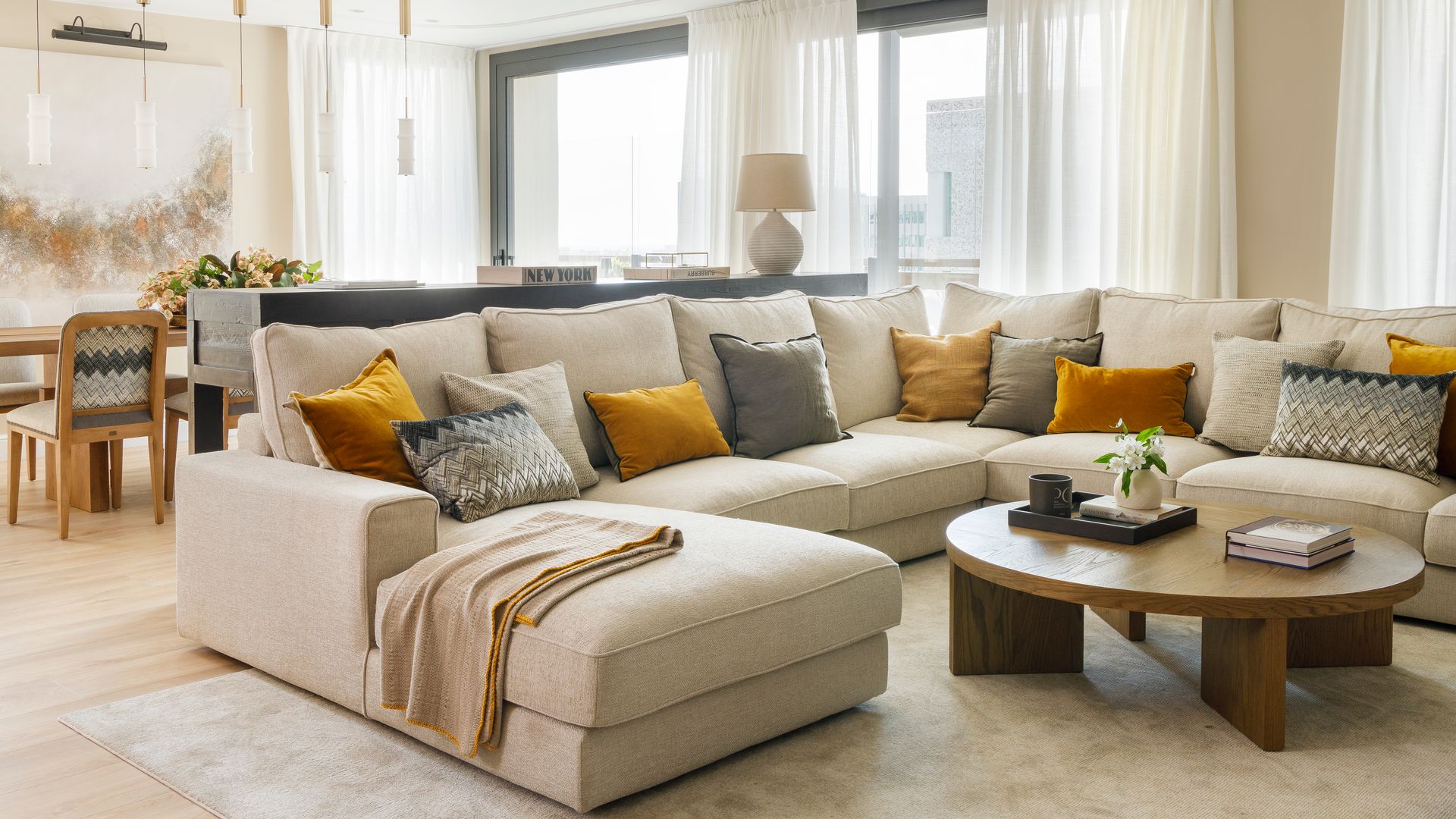 Salón con sofá XL y chaise longue en uno de sus lados, mesa de centro de madera redonda, alfombra, muchos cojines lisos en color mostaza y con motivos geométricos, manta en el sofá