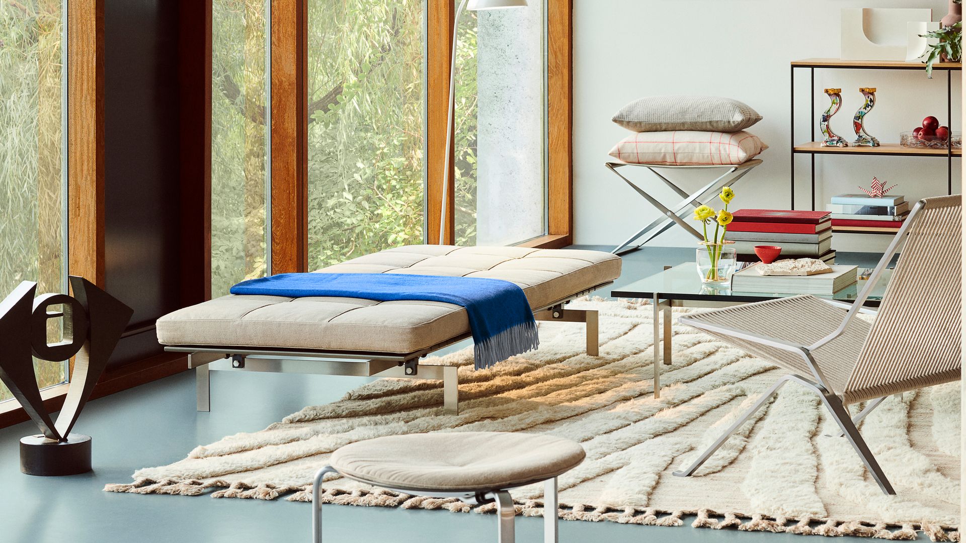 Salón moderno sin sofá con daybed blanca, alfombra beige, gran ventanal, estantería baja, silla trenzada, cojines, cuadros en la pared 