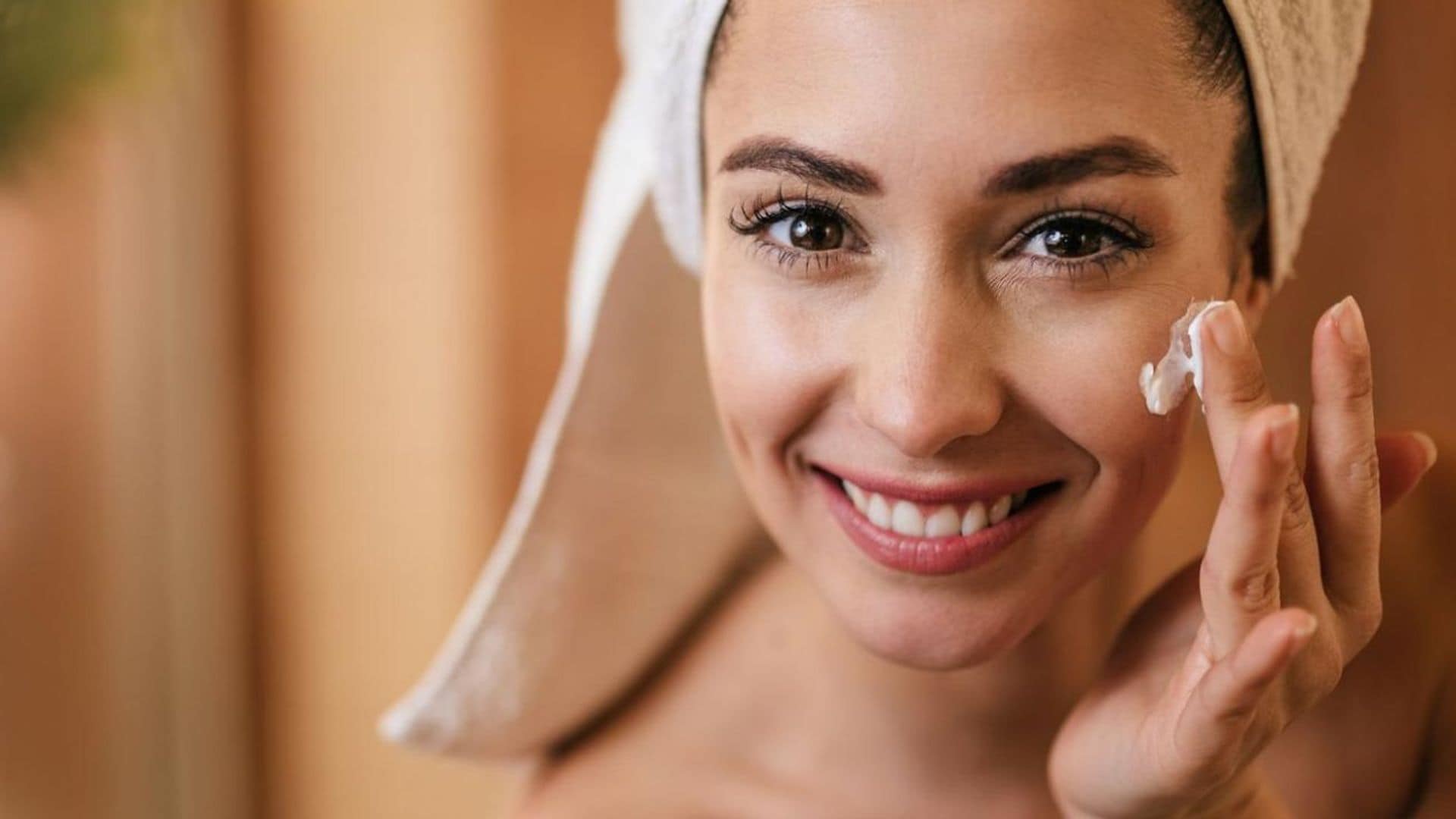 Mujer sonriente a la cámara recién salida de la ducha con toalla en el cabello, echándose crema en la cara.
