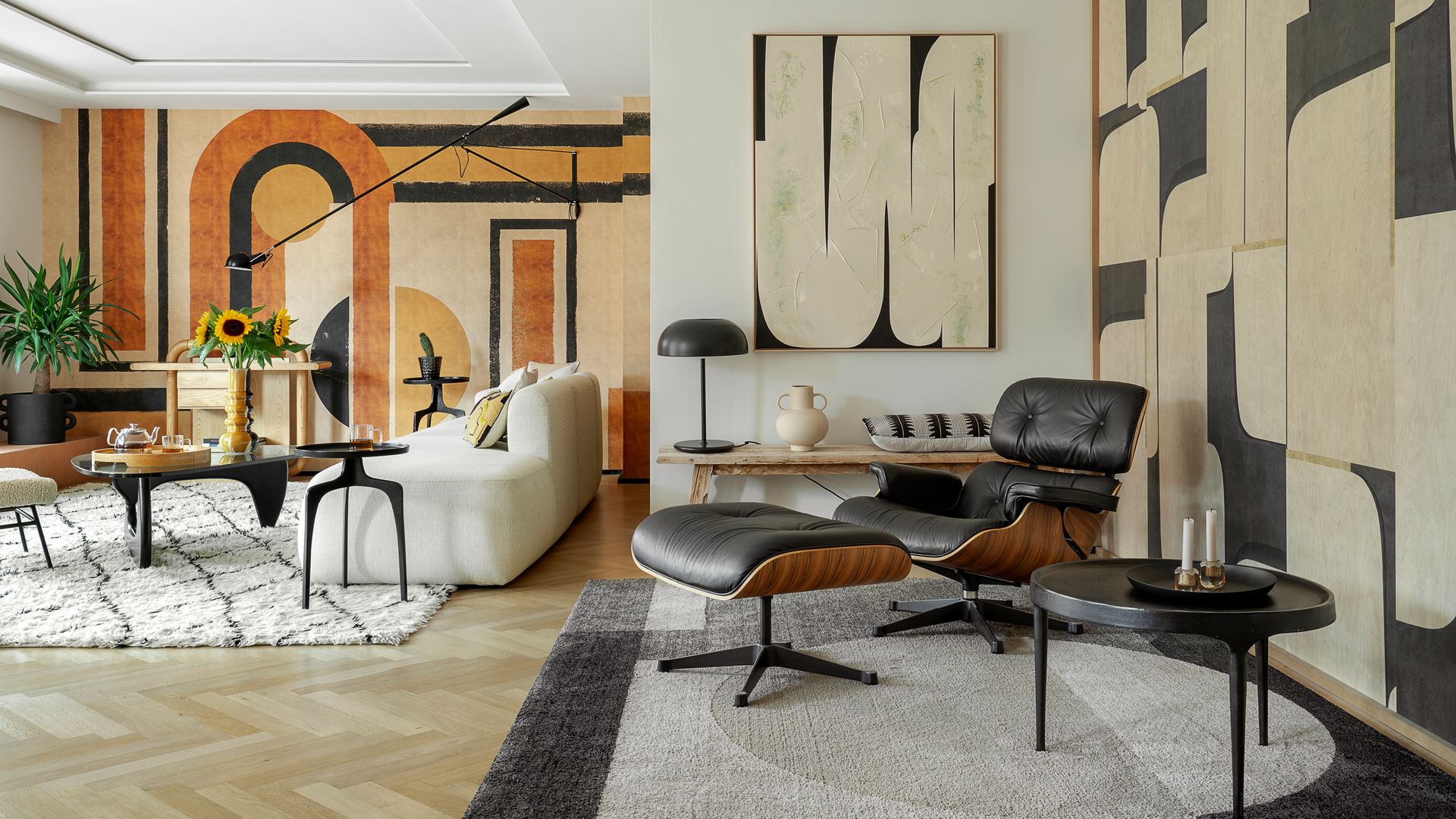 Salón con rincón de lectura, paredes con papel pintado con motivos geométricos y paneles, alfombras, chaise longue Eames, sofá blanco, mesa negra, jarrón con girasoles