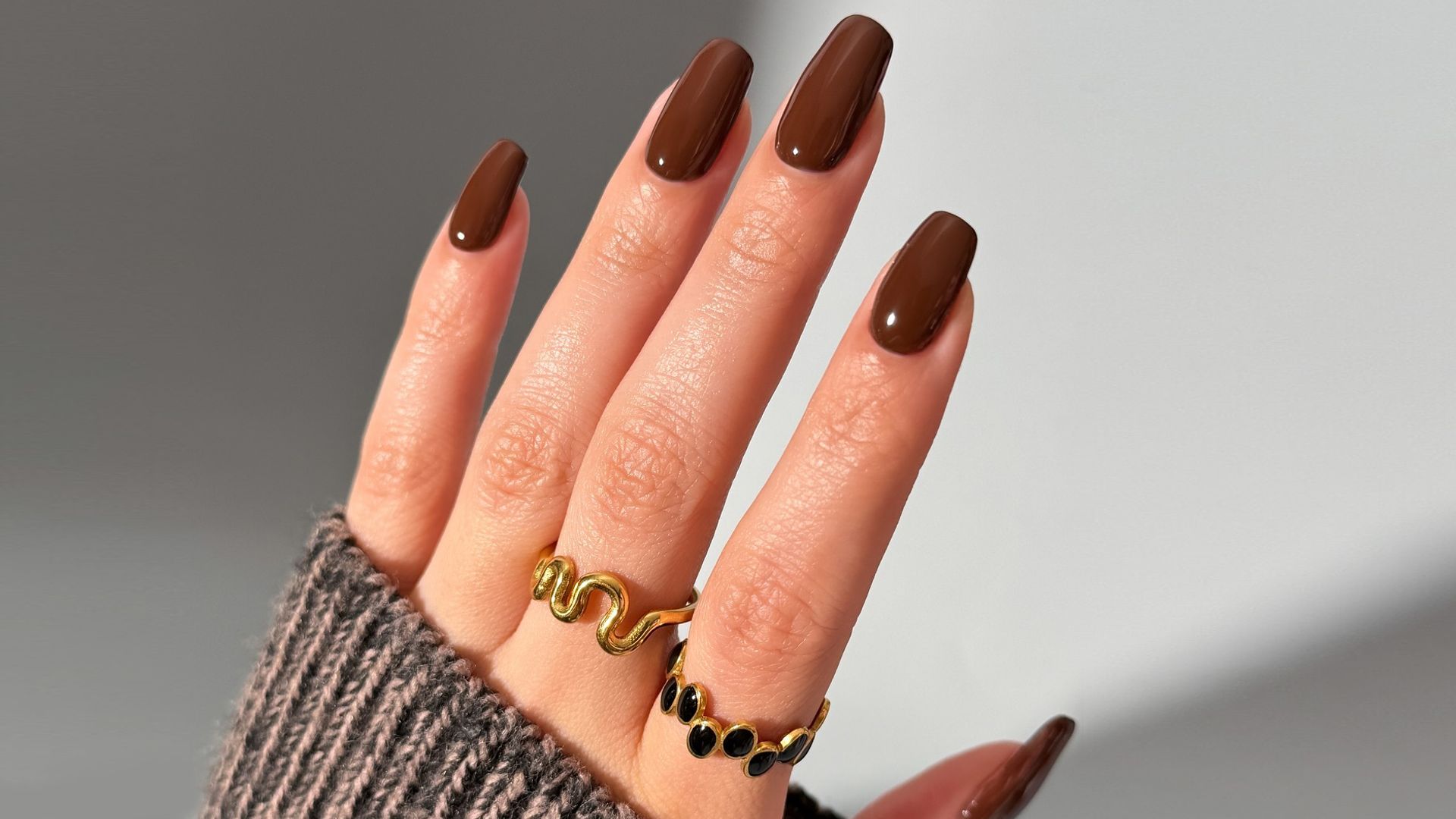 Mano con uñas marrones, jersey de punto marrón y dos anillos dorados