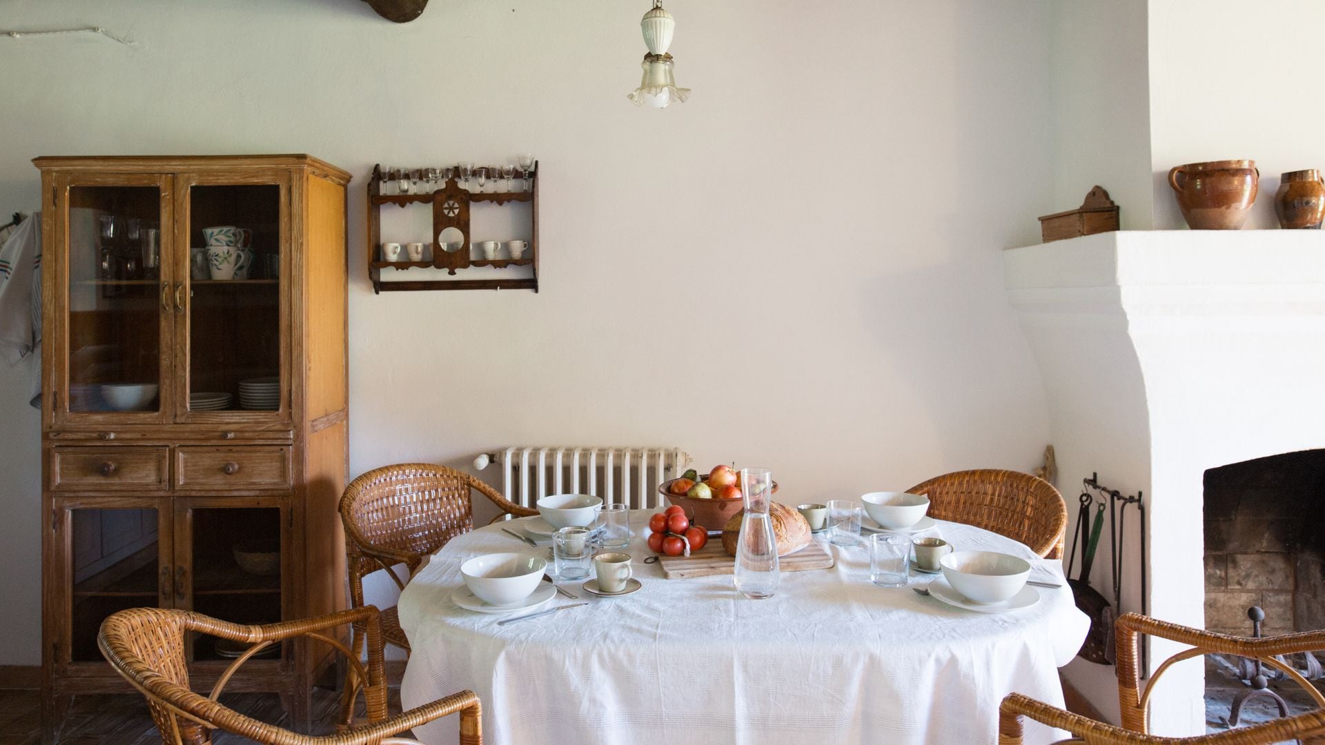 Comedor con chimenea, mesa redonda con mantel blanco, sillas de mimbre, vigas en el techo, vitrina alacena, platero de madera