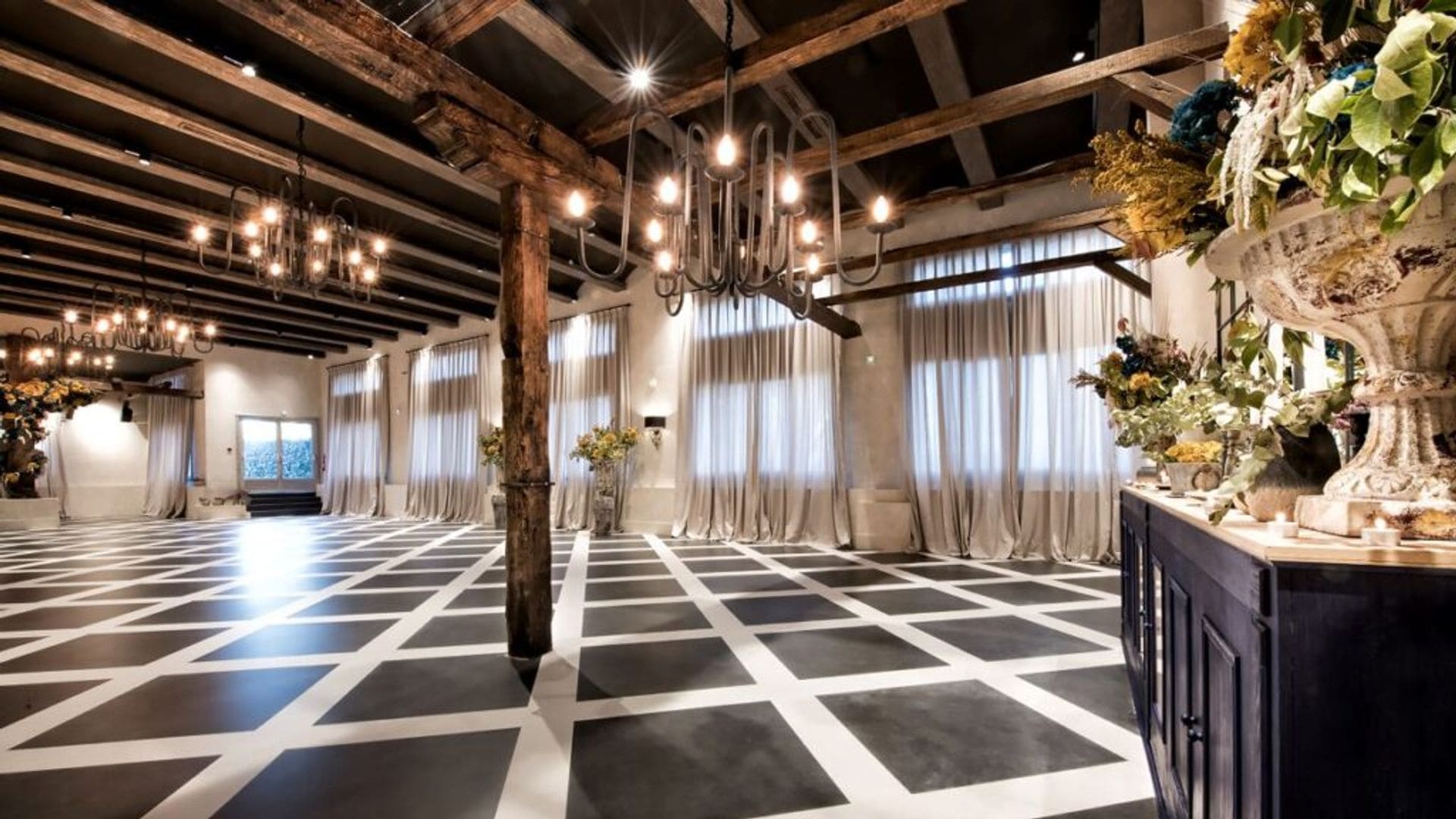 Salón con suelo en blanco y negro, vigas de madera, columnas de madera, lámparas de techo, cortinas, jarrón con flores, aparador 