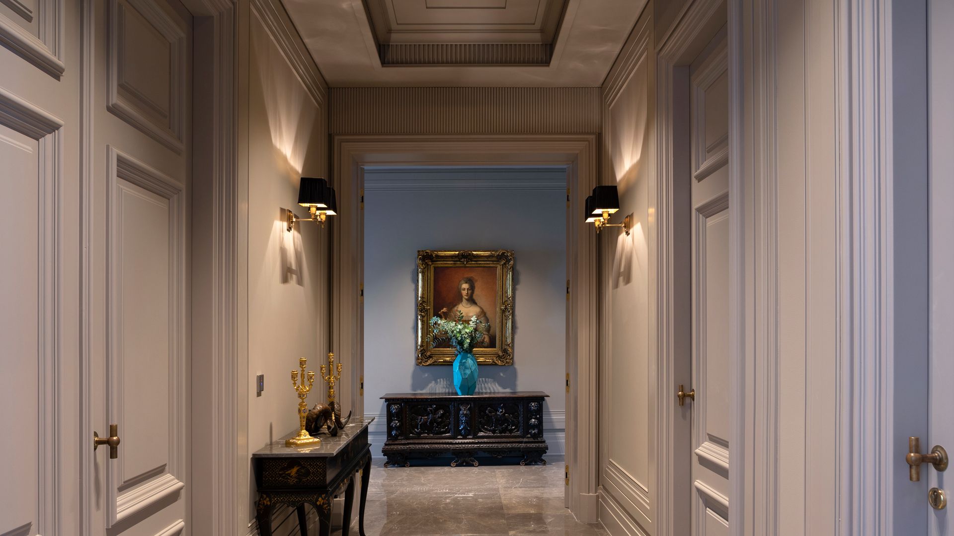 Pasillo con paredes y techos gris con molduras, suelo de mármol, focos empotrados, apliques de pared, consola en un lado y mueble negro al final con cuadro
