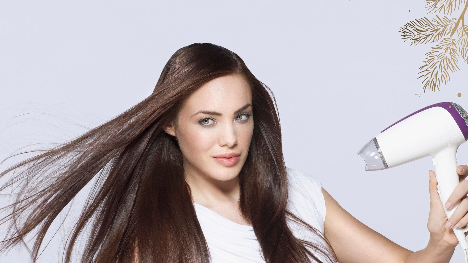 Mujer con cabello castaño y largo con un secador en la mano. Melena lisa, con camiseta blanca.