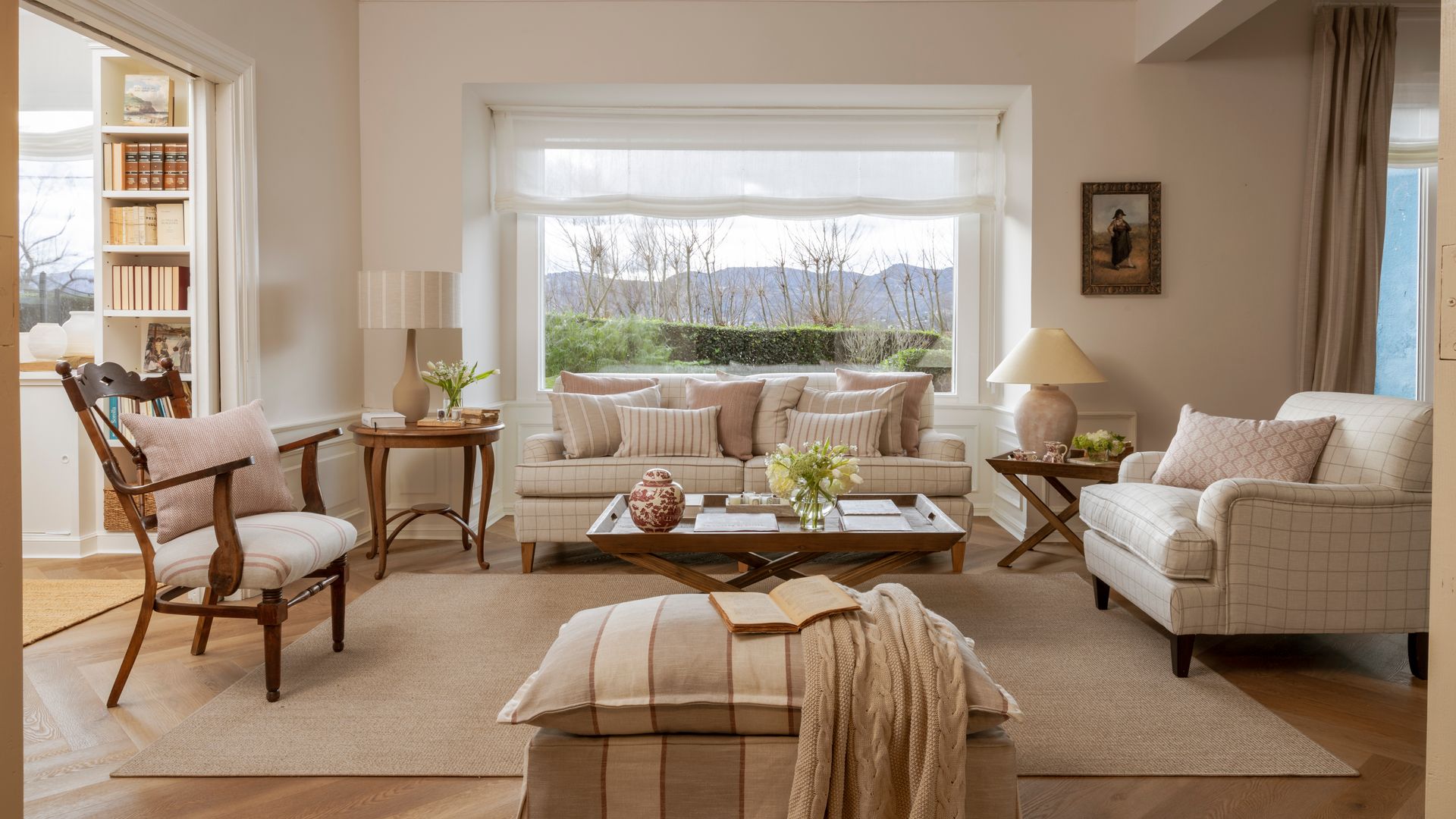 Salón con vigas en el techo, gran ventanal con estor blanco, alfombra, mesa de centro de madera, sofás blancos, sillas, sillón de cuadros beiges, cojines, puf