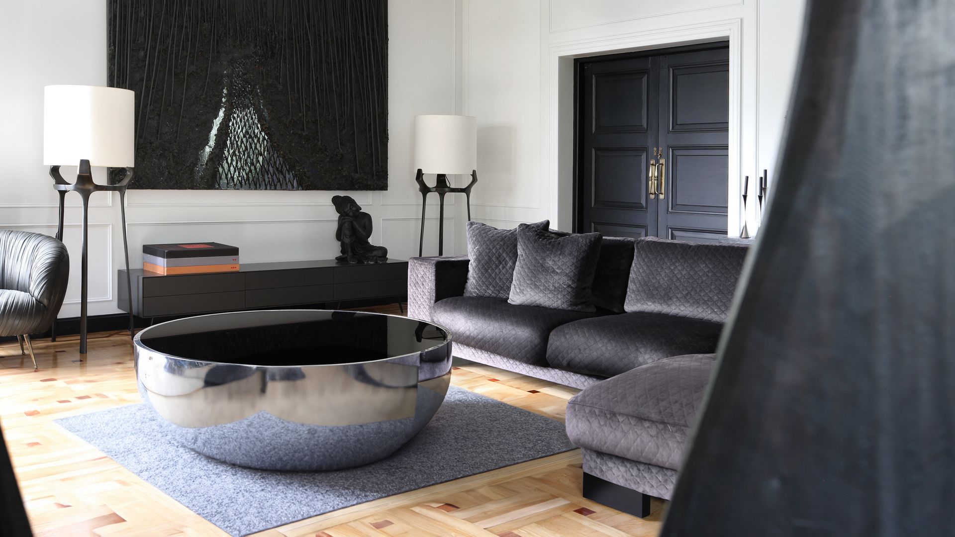 Salón con sofá de terciopelo negro, cuadro negro sobre una pared blanca, dos lámparas de pie, puerta negra, aparador bajo, mesa de centro redonda