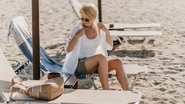 Mujer de 50 años en la tumbona de una playa, colocándose una camisa blanca sobre una camiseta de tirantes blanca. Está sentada, con una toalla y una bolsa de playa en la tumbona de delante.