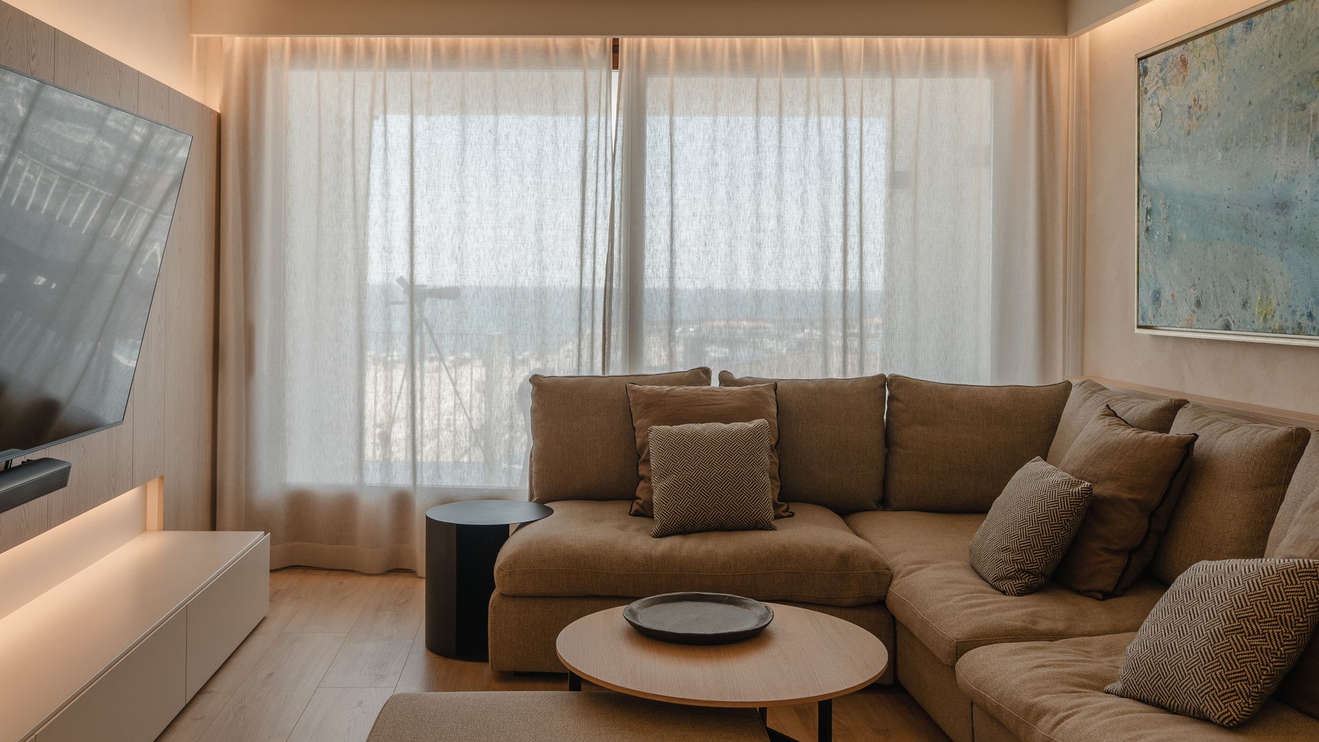 Salón en tonos beige con sofá en marrón claro, television colgada de la pared, cortinas claras, cuadro en la pared del sofá 