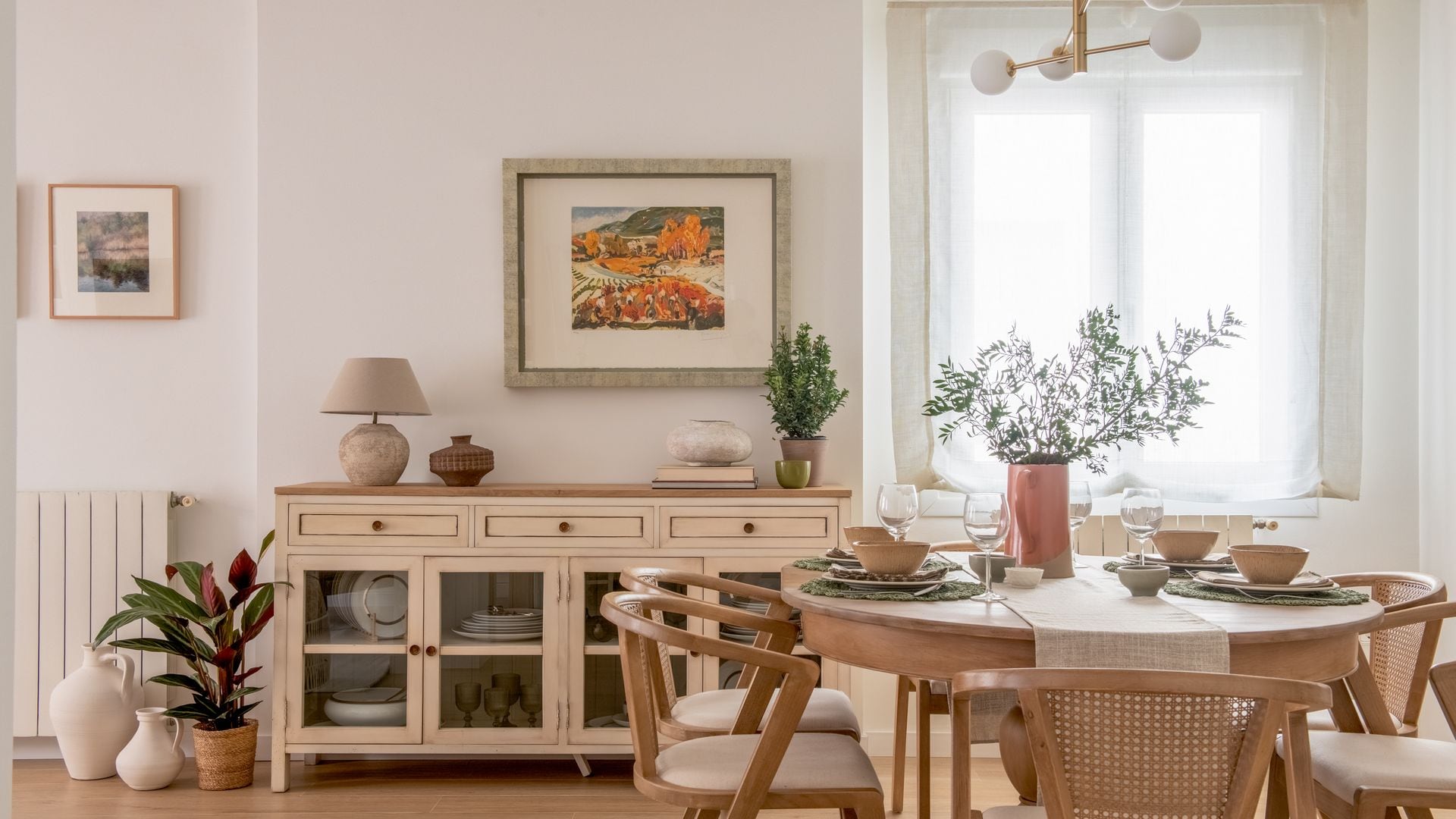 Comedor con mesa redonda de madera, aparador con vitrina en la parte inferior, lámpara de techo, jarrones, mesa con manteles individuales y camino de mesa, estor blanco, radiador