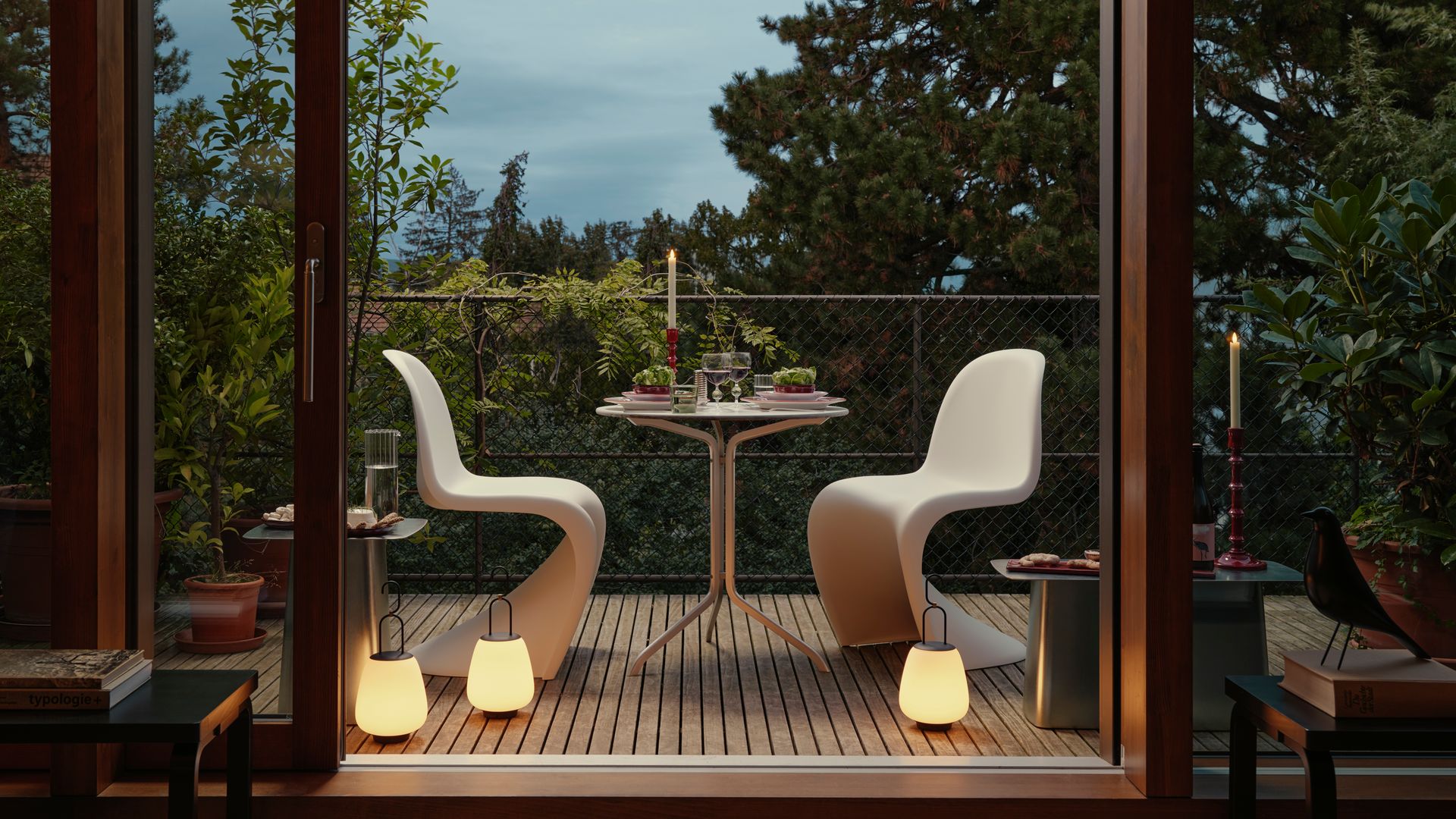 Terraza pequeña con suelo de madera, lámparas portátiles en el suelo, sillas Panton blancas, mesa de comedor mini