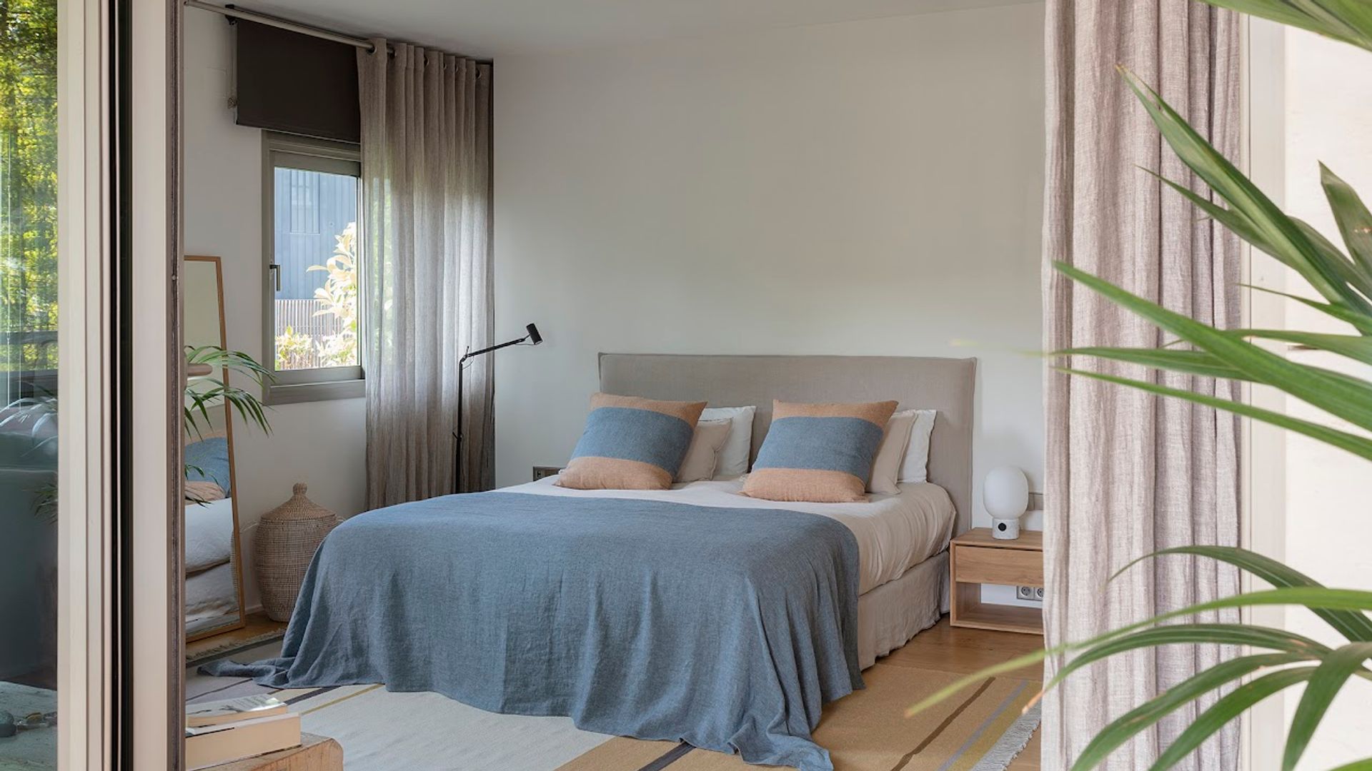 Dormitorio con cabecero tapizado, alfombra, ropa de cama en color azul, mesitas de noche de madera, lámpara de pie, espejo, cortinas y estores