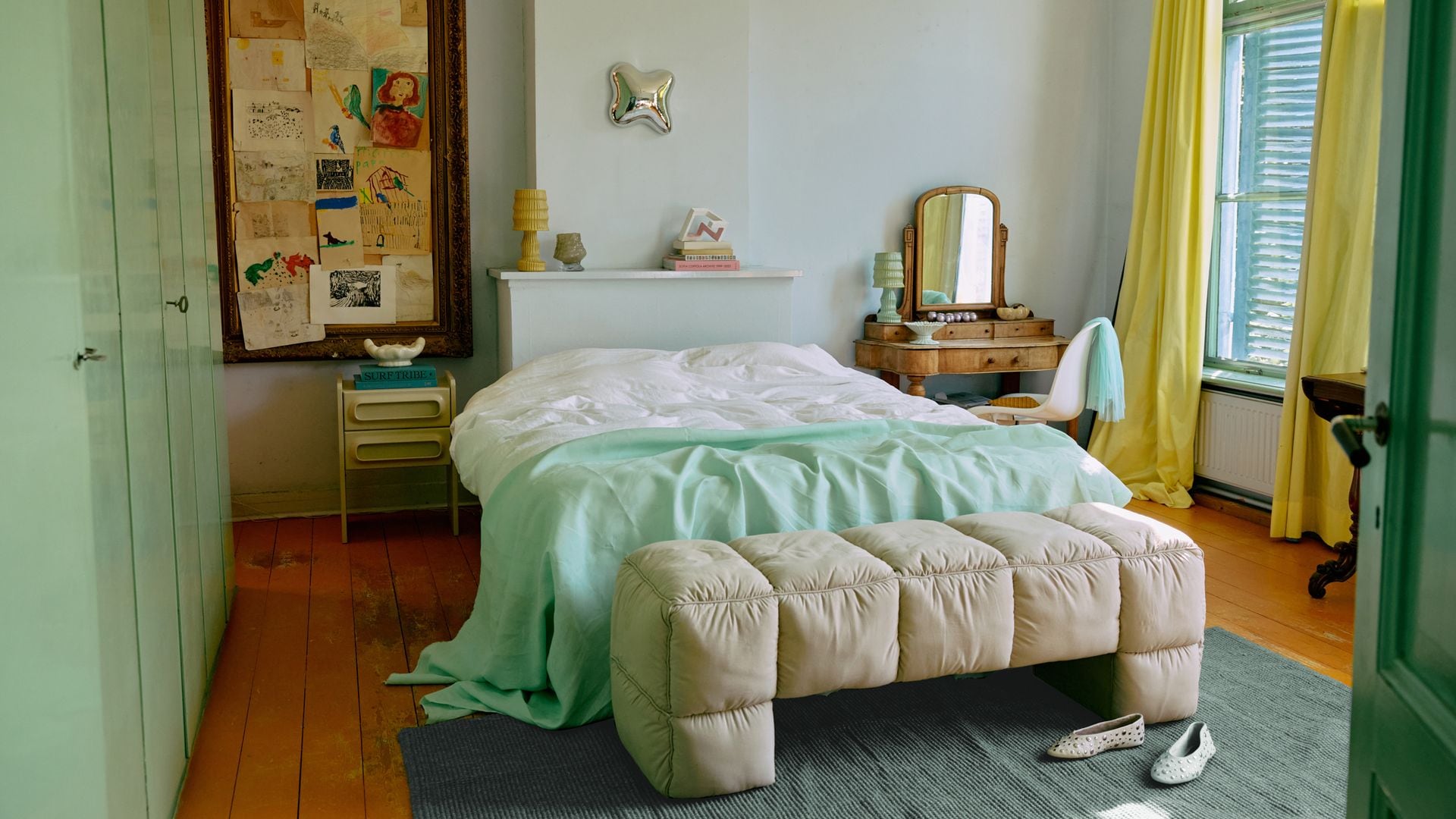 Dormitorio con banqueta tapizada a los pies de la cama, cortinas amarillas, armario verde menta, plaid verde menta, tocador con espejo, alfombra, marco de cuadro con dibujos y papeles