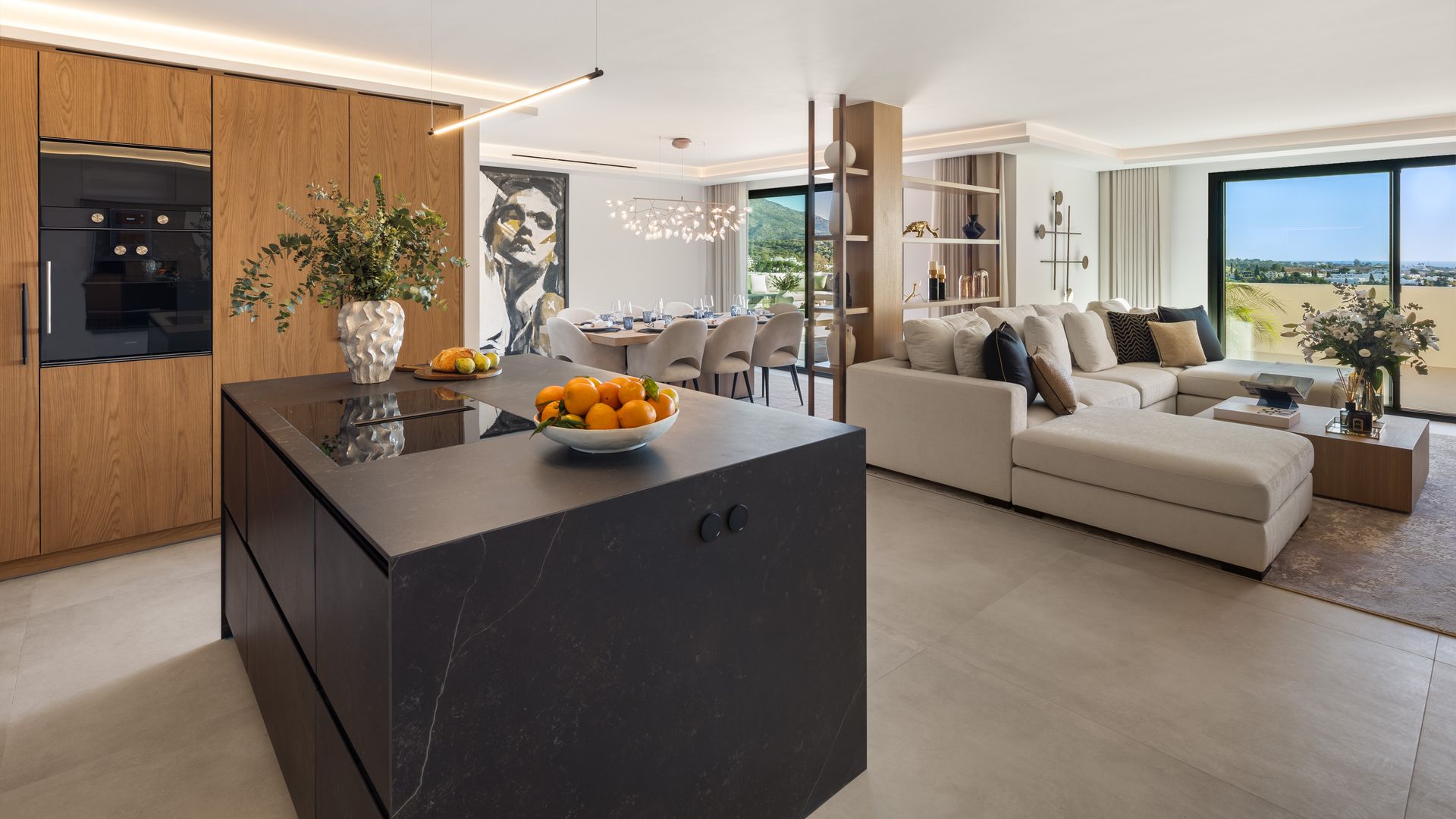 Cocina con isla abierta al salón y comunicada con el comedor, isla negra, cuadro en la pared del comedor, sofá con chaise longue, armario de madera con electrodomésticos negros