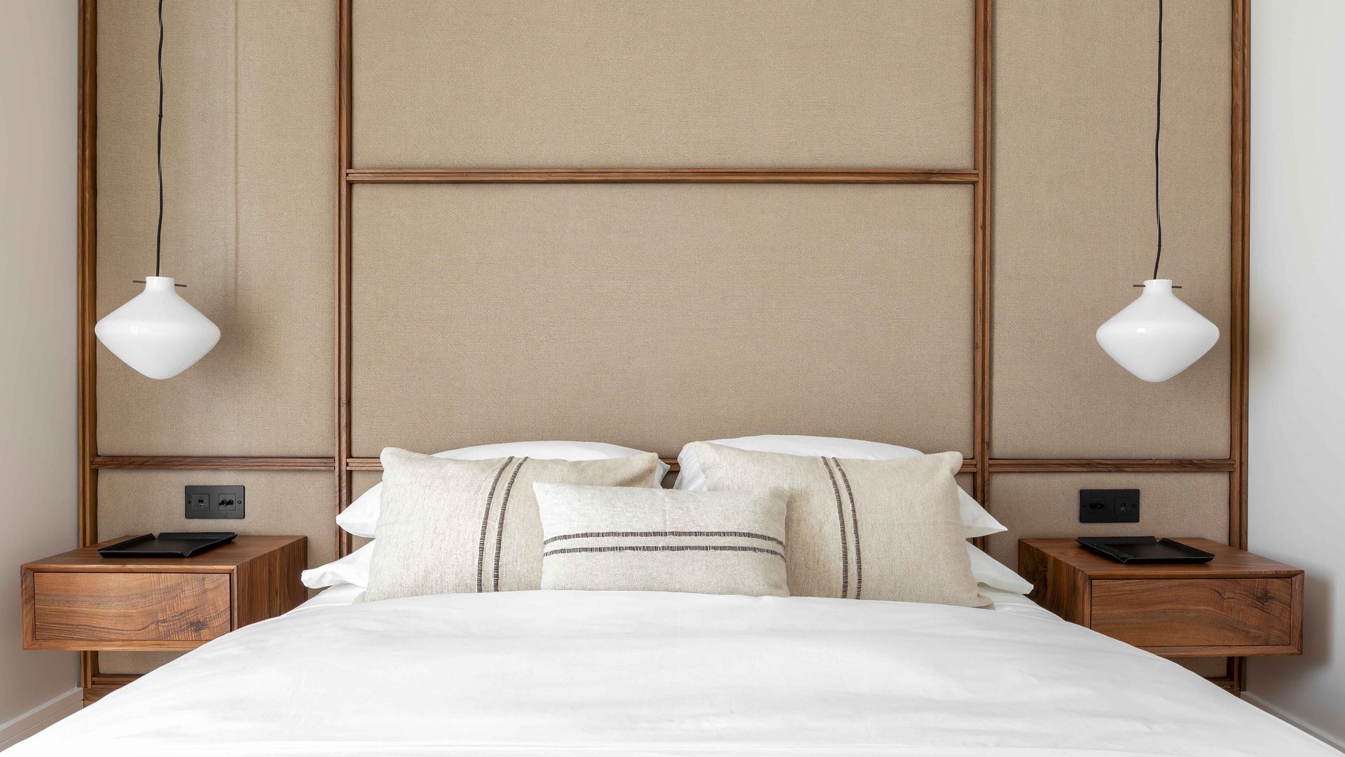 Dormitorio con pared del cabecero revestida con paneles tapizados en beige con marco de madera, mesitas de noche de madera suspendidas, cojines con ribete, ropa de cama blanca, plaid de cuadros marrones y blancos, lámparas de techo blancas a ambos lados de la cama