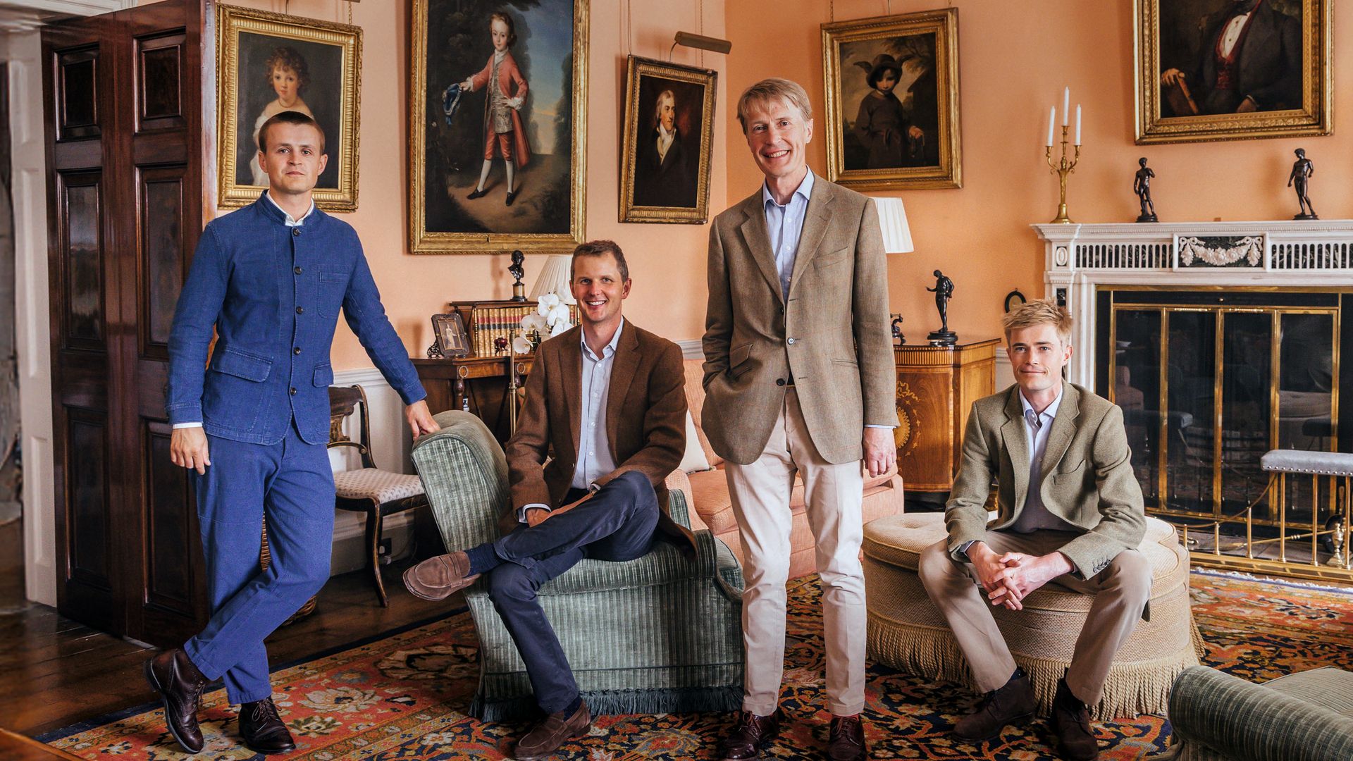 Cuatro hombres en trajes elegantes posan en una sala decorada con cuadros y una chimenea.