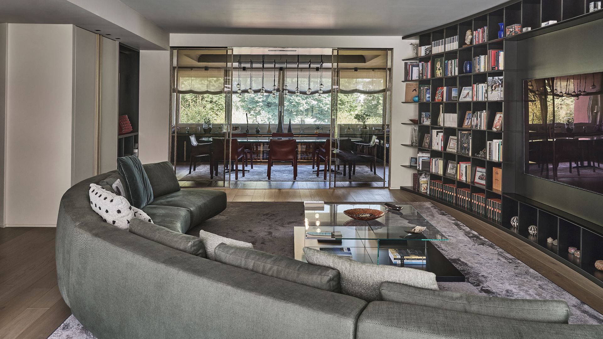 Salón comedor con una gran librería curva negra, sofá gris curvo, televisión en la librería, alfombra gris