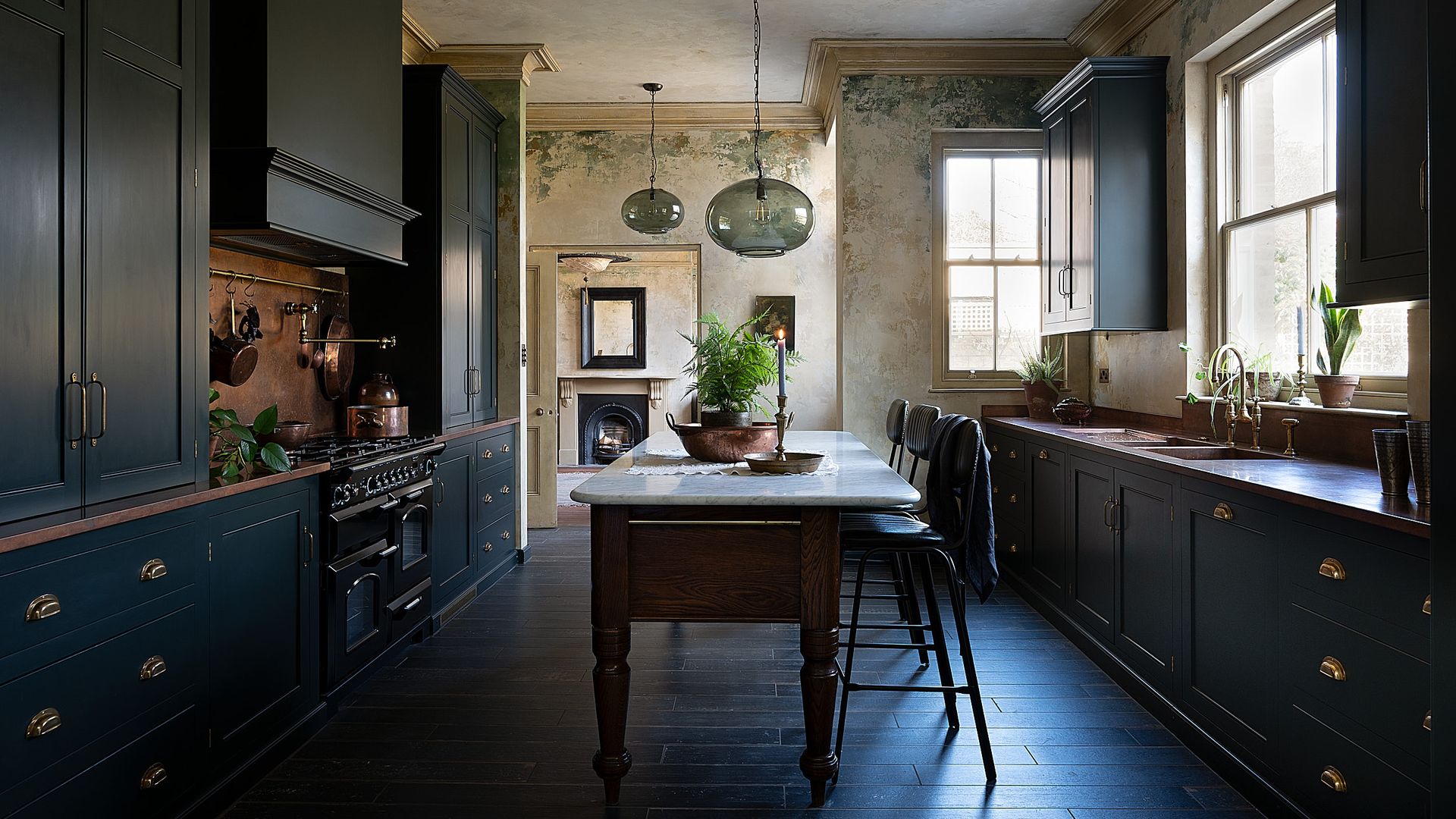 Cocina de estilo inglés con los muebles en azul oscuro, el suelo de madera teñida, mesa de madera antigua con el sobre de mármol, taburetes, vela encendida, lámpara de techo de cristal 