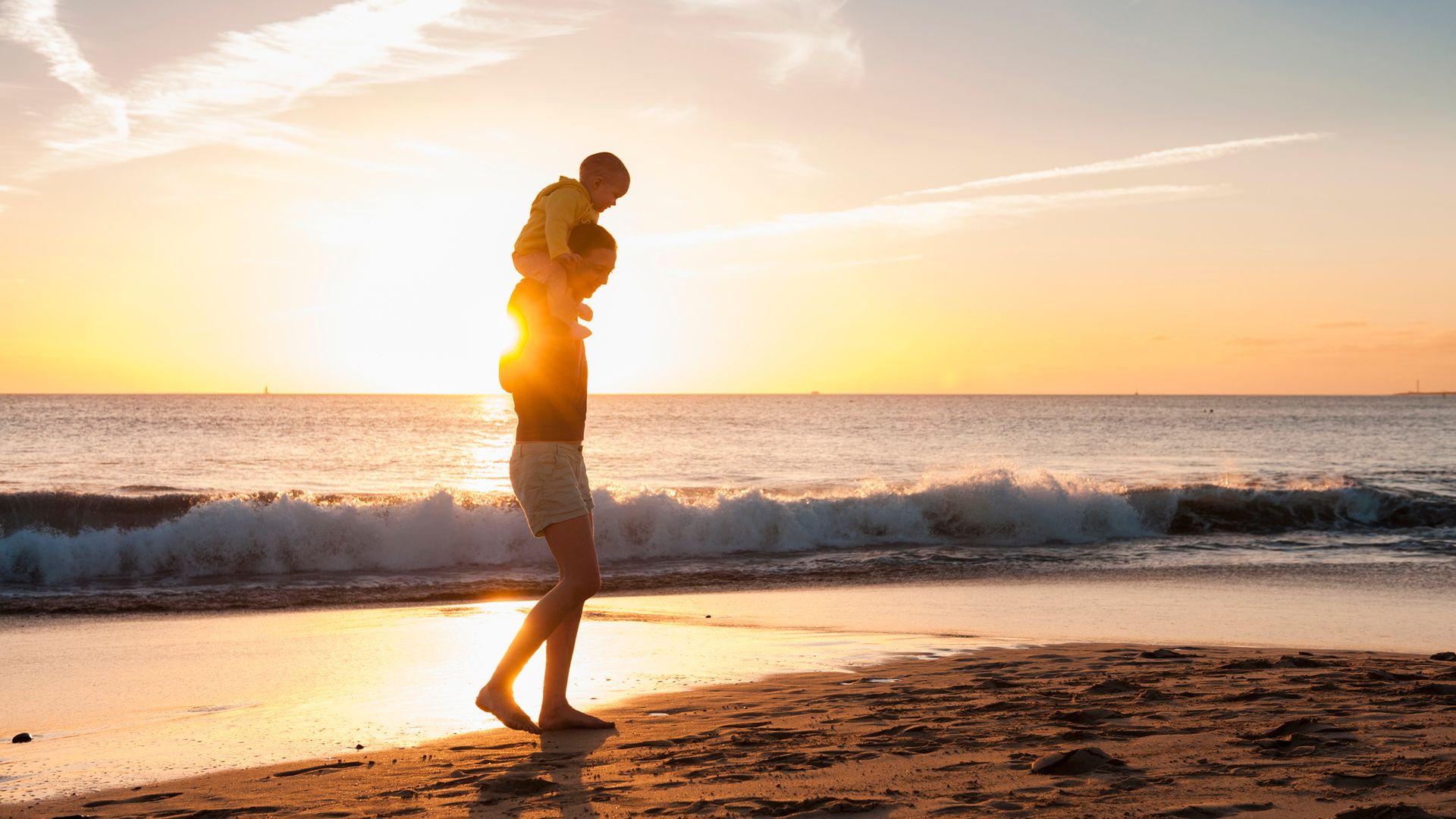 Madre con su bebé a los hombros paseando por la playa al atardecer, mientras ambos sonríen.