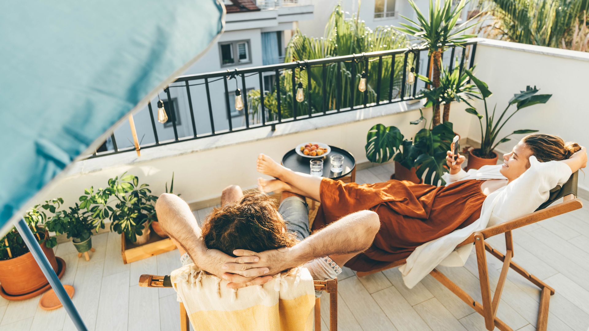 Dos mujeres relajadas en una terraza urbana, muy bonita decorada y con muchas plantas. tienen tumbonas, una mesa y están tomando un aperitivo.
