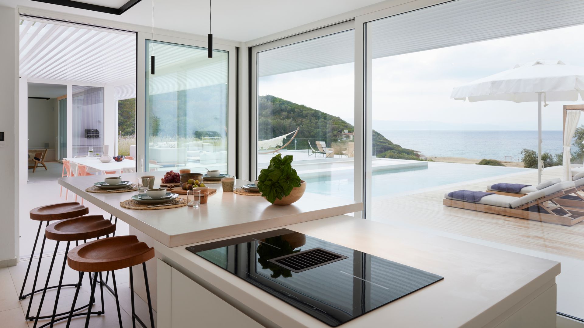 Cocina con isla con zona de fuegos y zona de comedor con taburetes altos, grandes ventanales que conecta directamente con la piscina 