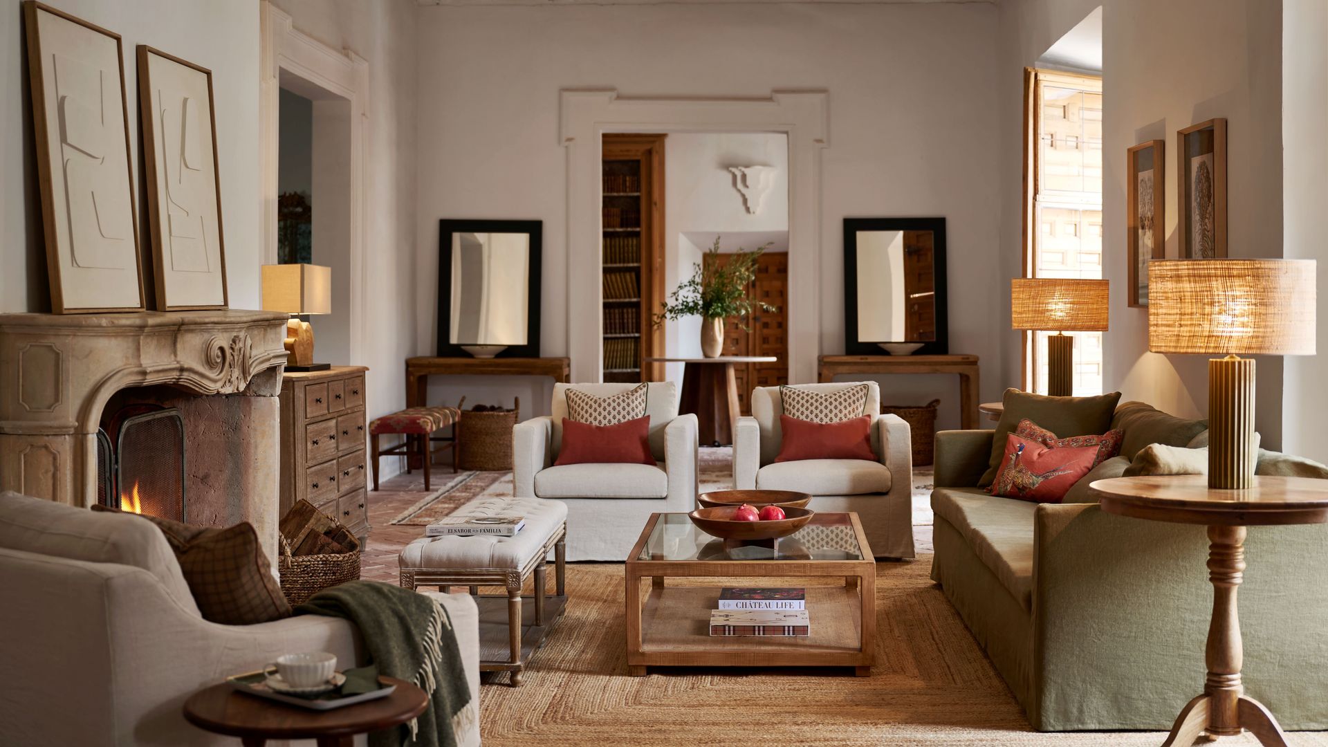 Salón con chimenea, con cuadros sobre la chimenea, alfombra de fibras vegetales, mesa de cristal y madera, sofá verde, butacas blancas con cojines, taburete de madera con taza de café, aparador alto de madera tipo archivador, lámparas de sobremesa encendidas 