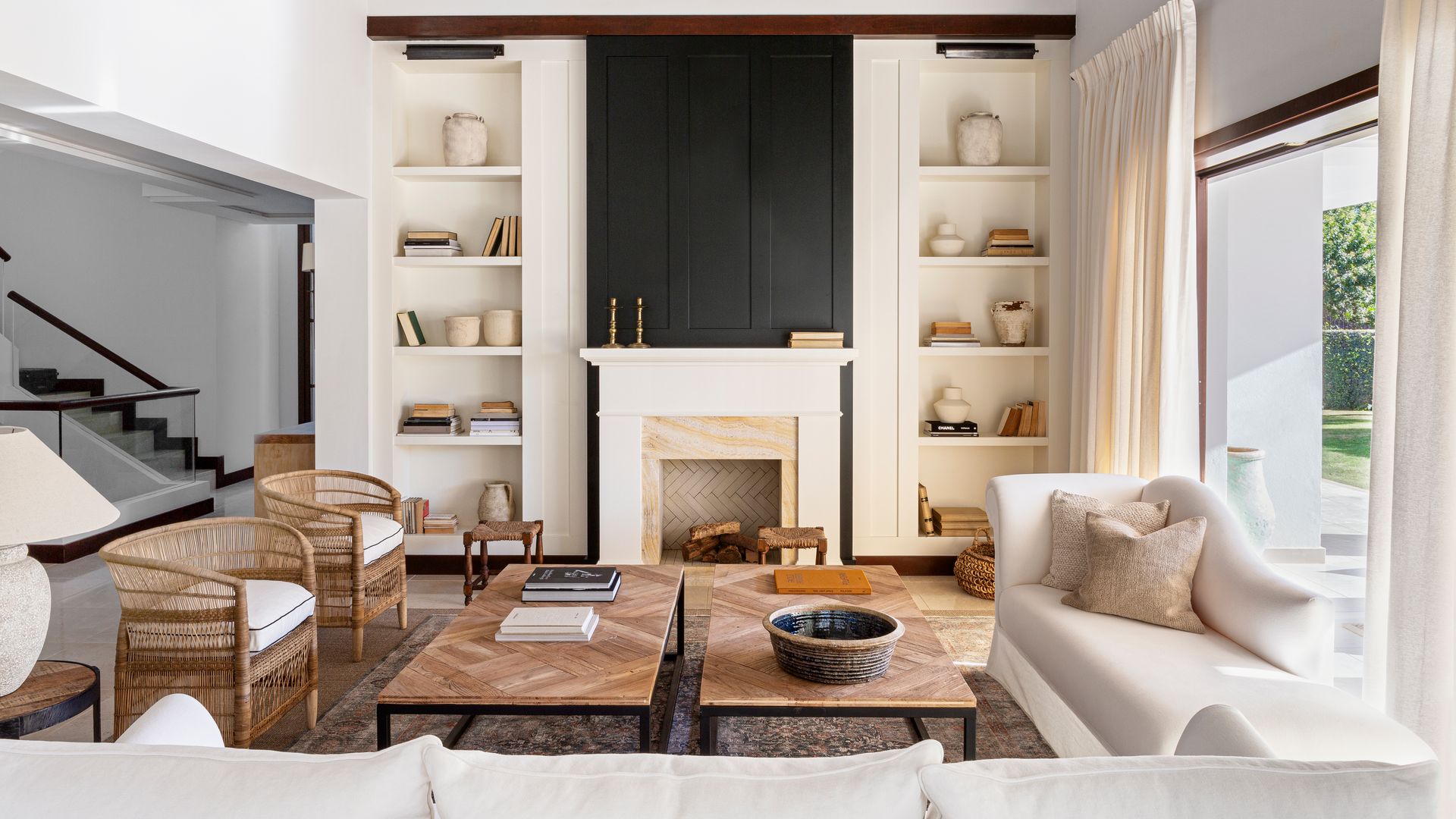 Salón con chimenea, con estanterías a los lados, cortinas, sofás blancos, butacas de mimbre, mesas de centro 